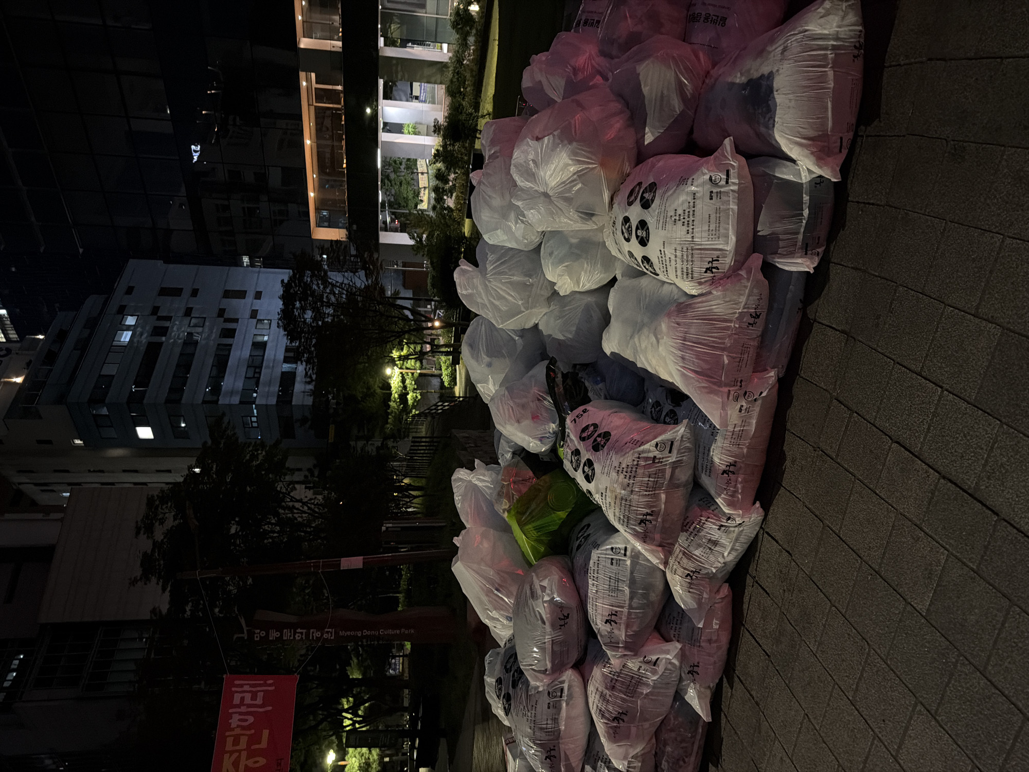 The image shows a large pile of trash bags on a brick sidewalk, likely for collection. The bags are mostly white and translucent, with some having printed Korean text and symbols on them. A single green bag is visible within the pile. In the background, there are buildings, including a taller one with illuminated windows. A sign indicates that the location is Myeong Dong Culture Park, a public space in Seoul, South Korea. The scene appears to be at night, with artificial lighting illuminating the area.