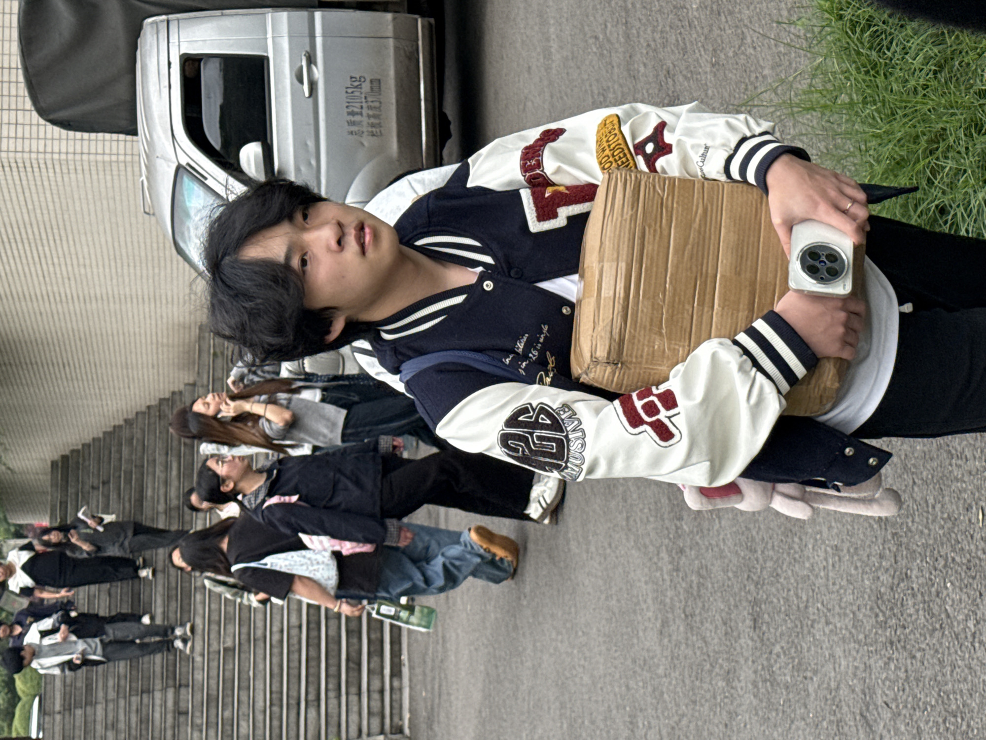 A young Asian person in a varsity jacket and black pants stands facing the viewer and holding a cardboard box, a white smartphone, and a white stuffed toy. The person is outside near a parked silver van with Chinese writing on the side. In the background, a group of people stands on stairs. Vegetation grows on the upper right. The location is presumed to be Jiangjin, China.