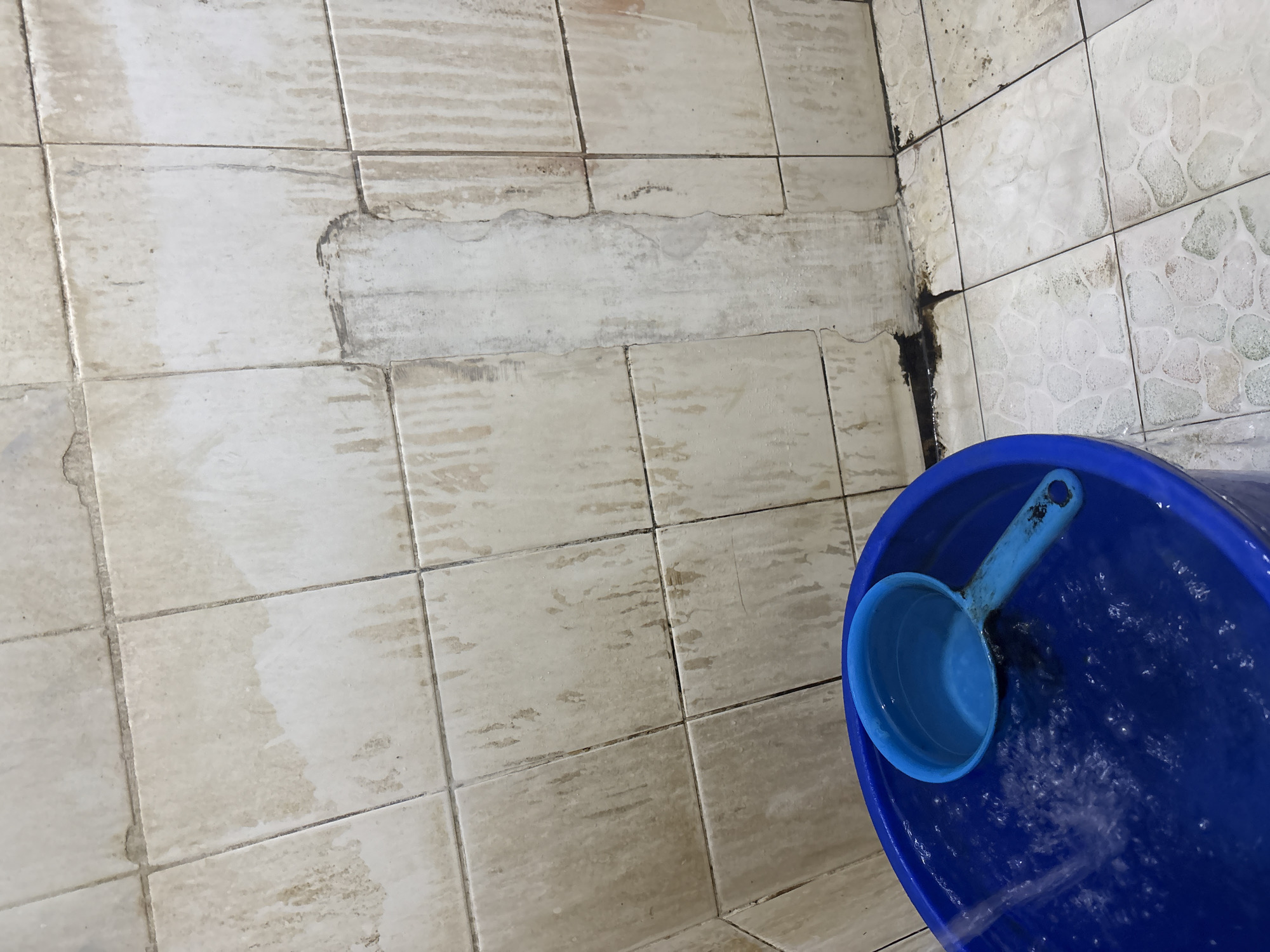 The image depicts a close-up view of a corner of a bathroom in Palimanan, Indonesia. The walls are tiled with light-colored square tiles, which appear to be stained and discolored with streaks of grime, suggesting a lack of recent cleaning. In the foreground, a large blue plastic bucket is partially filled with water. A blue plastic ladle rests inside the bucket. Water is actively flowing into the bucket, creating ripples on the surface. The floor is also tiled, with a pattern of lighter-colored, irregular shapes on a white background, also showing signs of staining. The overall impression is of a functional but neglected bathroom space.