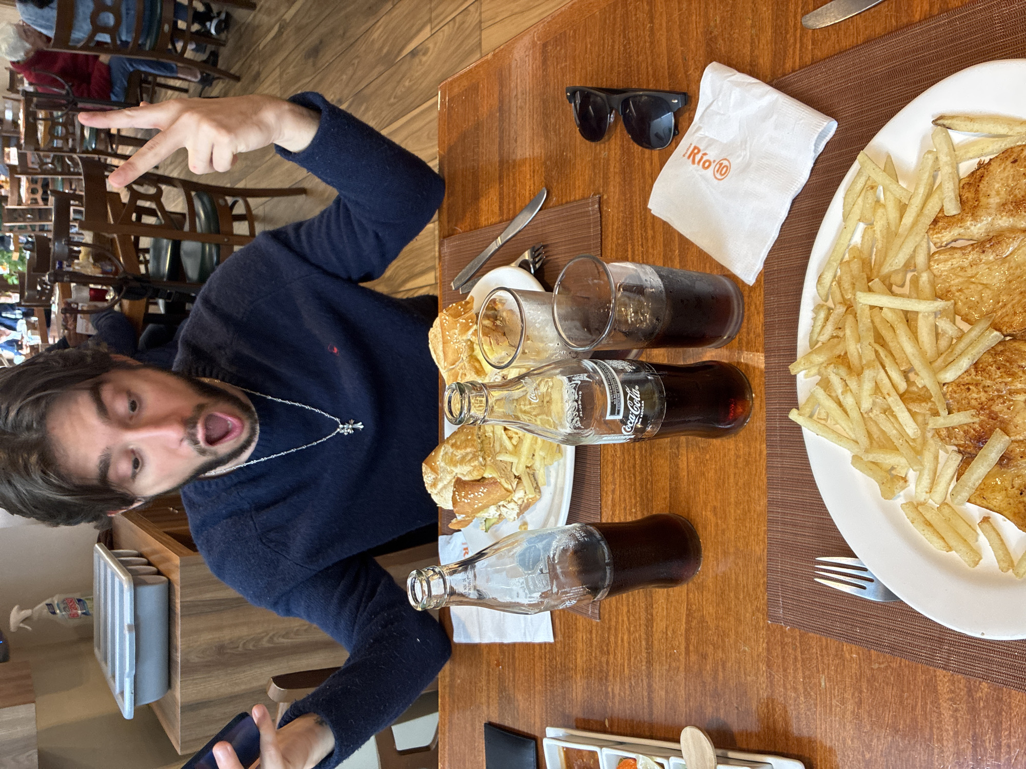 A man with dark hair and a beard, wearing a dark blue sweater and a silver necklace, is seated at a wooden table in a restaurant. He is holding a smartphone in his left hand and has his right hand raised with two fingers extended in a peace sign. His mouth is open in a surprised expression. In front of him are two bottles of Coca-Cola, two glasses of dark liquid, a plate of food with a sandwich and french fries, a white napkin with "Rio" printed on it, and a pair of black sunglasses. A fork and knife are also visible on the table. The background shows other patrons and tables in the restaurant. The restaurant appears to be in Cuautitlán Izcalli, Mexico.