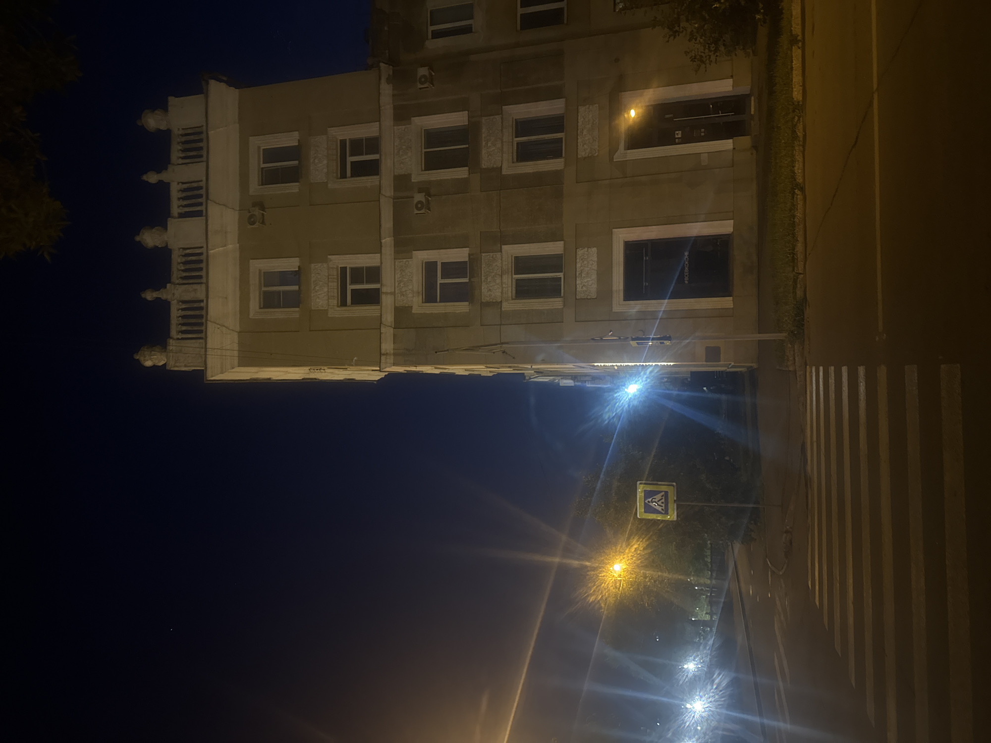The image depicts a nighttime street scene in Magnitogorsk, Russia, featuring a multi-story building and a pedestrian crossing. The building is tall and has a light-colored facade with several windows and what appears to be decorative molding. The top of the building is adorned with ornate balustrades and finials. Streetlights cast a yellow and white glow, creating starburst effects due to the camera's aperture. A pedestrian crossing is clearly visible in the foreground, marked by white stripes on the asphalt. A yellow pedestrian crossing sign is mounted on a pole to the left of the building, next to the crosswalk. The sky is dark blue, indicating nighttime, and there are a few faint lights in the distance. The overall impression is one of an urban environment at night, with artificial lighting illuminating the scene.