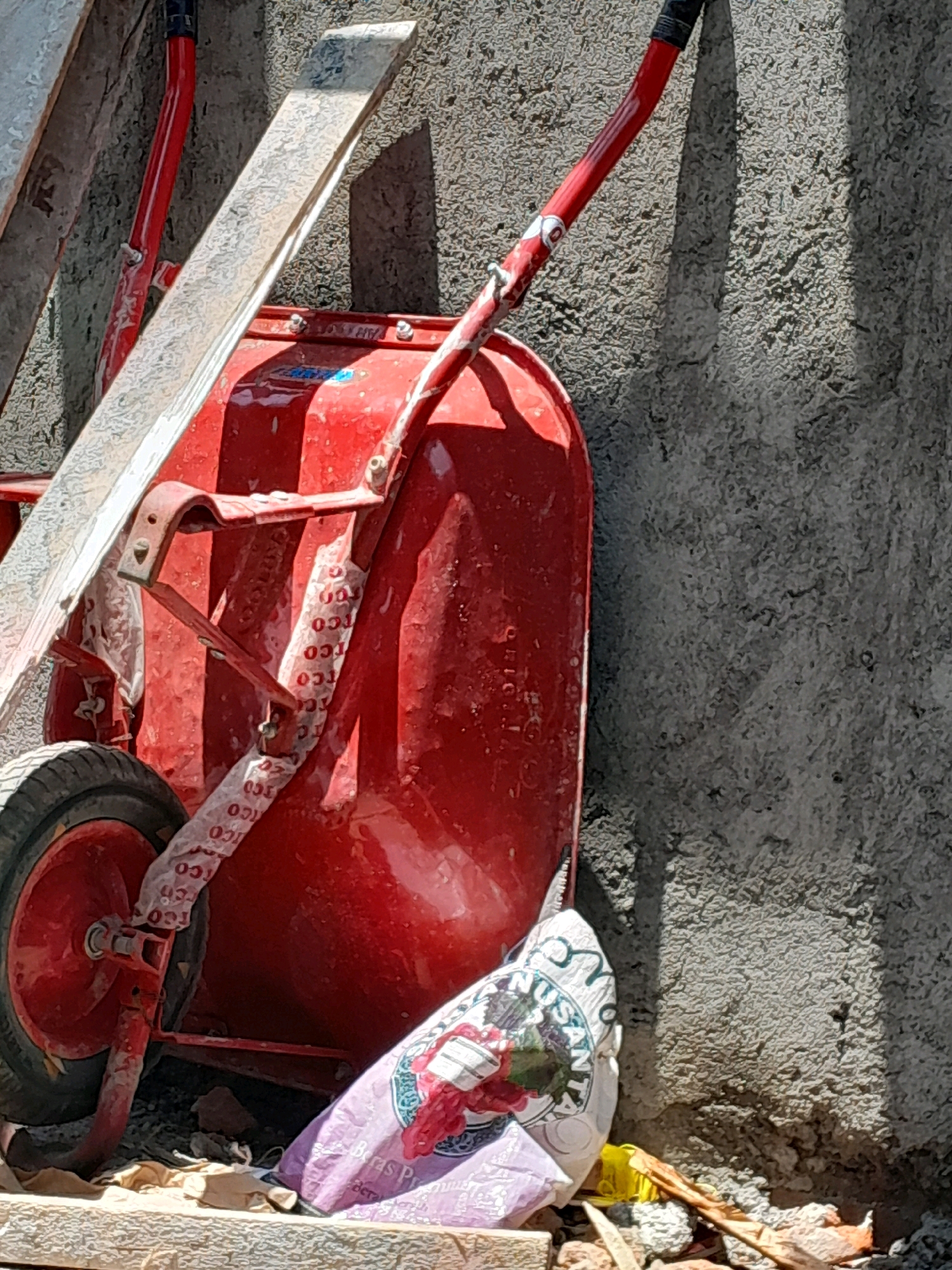 A red wheelbarrow with a black tire is leaning against a rough, textured concrete wall. The wheelbarrow is partially covered with white plastic sheeting that has red lettering. A section of what appears to be a wooden plank or ladder rests diagonally across the wheelbarrow. In the foreground, a purple bag with printed graphics is visible, along with some debris on the ground, including small rocks and pieces of wood. The scene appears to be outdoors, likely in an area under construction or storage. This image was captured in Karangduren, Indonesia.