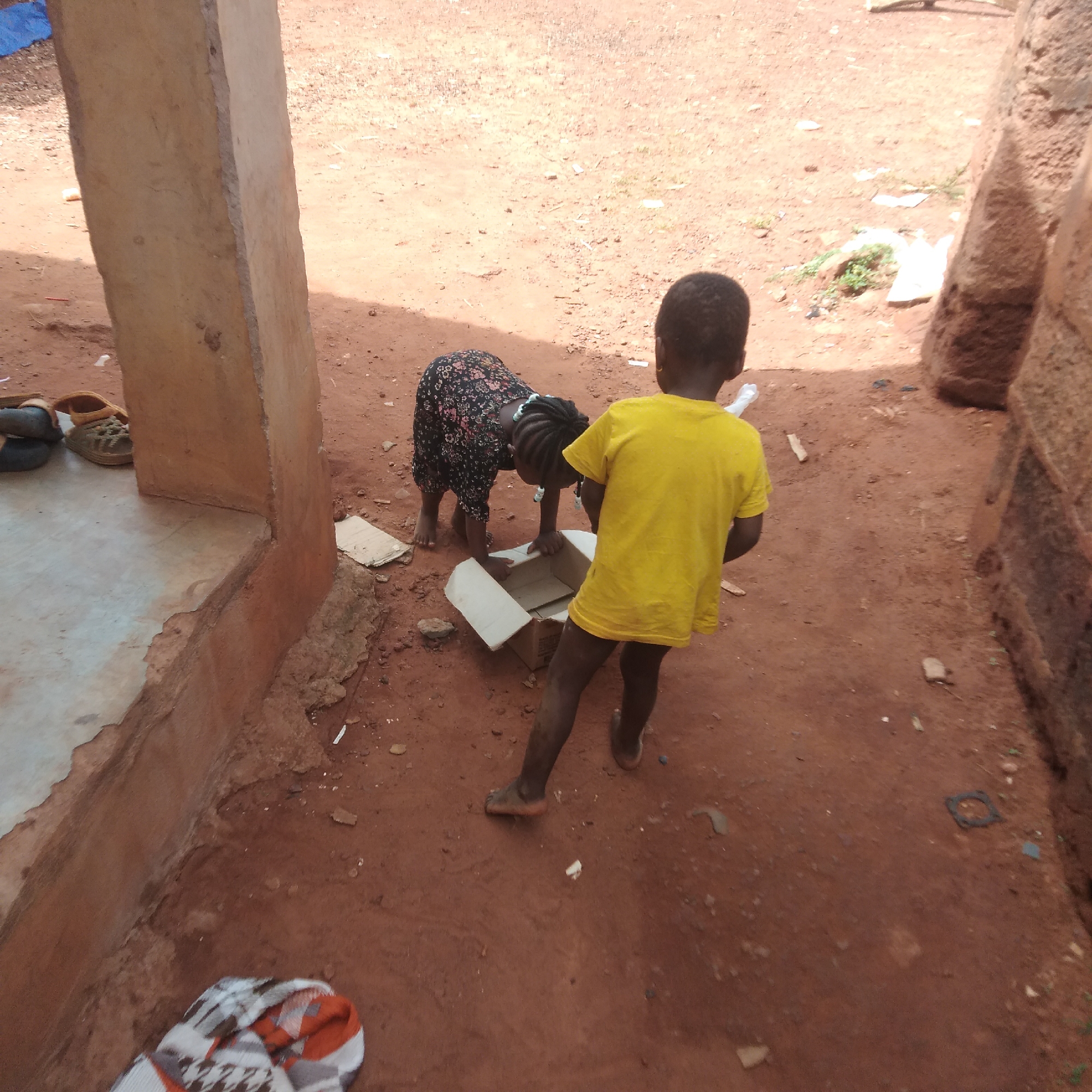 The image depicts two young children in Ejule, Nigeria, engaged in play near a structure made of concrete and reddish-brown brick. One child, wearing a yellow t-shirt, stands behind a partially open cardboard box, while the other, dressed in a dark floral print dress, leans over the box, seemingly looking inside. The ground is reddish-brown dirt with scattered debris. A pair of sandals are visible near the concrete structure. A blanket or cloth with a geometric pattern lies on the lower left edge of the frame. The scene is outdoors and appears to be a residential area.