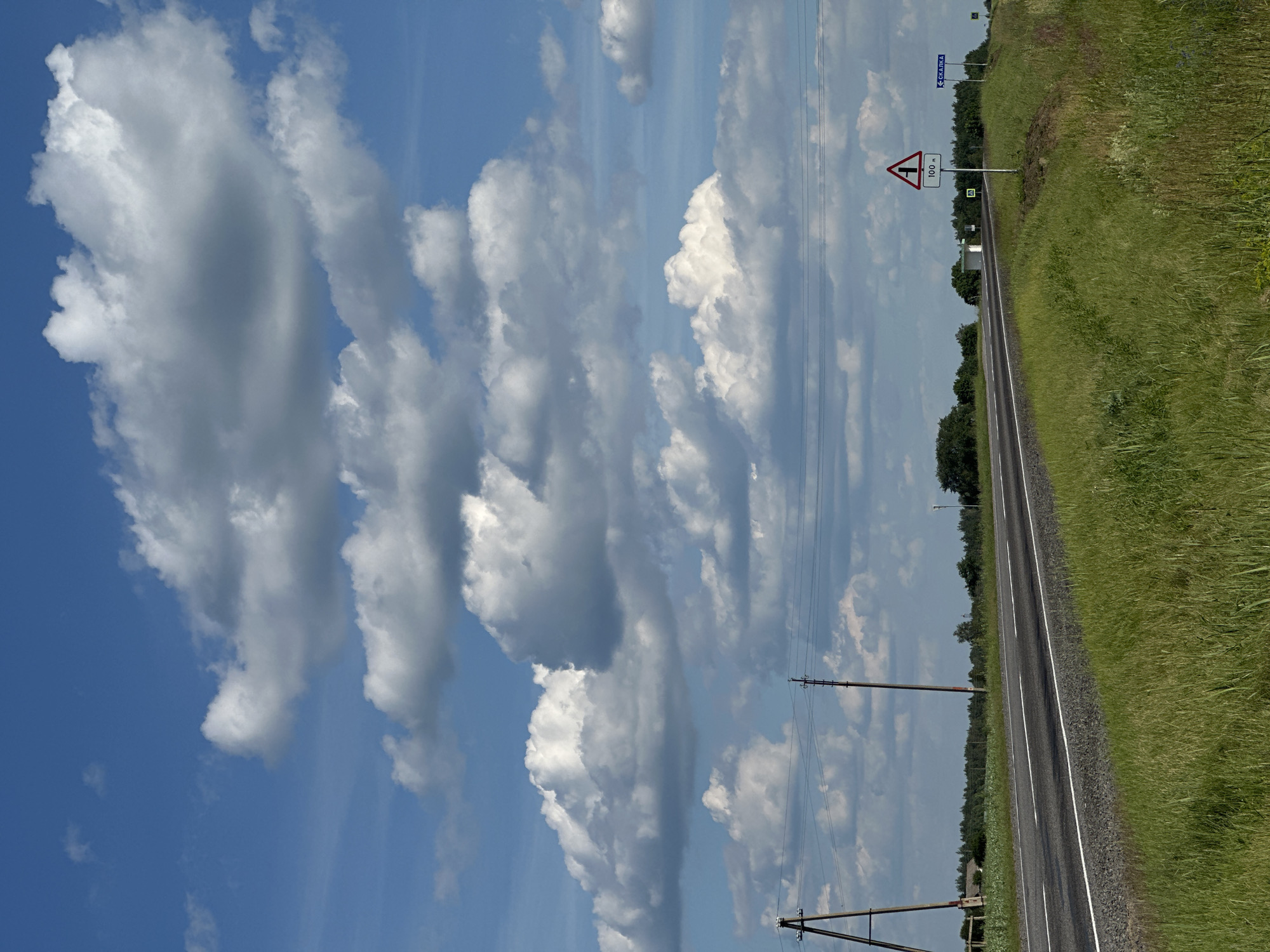 The image depicts a rural roadside scene in Belarus under a bright, partly cloudy sky. The perspective is from a low angle, looking up at the sky and along the road. In the foreground, a strip of lush green grass lines the right side of a paved road with a white stripe down the center. Beyond the road, a dense line of dark green trees forms the horizon.
Several power lines and utility poles traverse the scene, one prominently in the lower right quadrant, and others receding into the distance. A triangular road sign with a red border indicates a sharp turn ahead and reads "100 m" below the symbol. Another smaller, square green sign with a white symbol is visible further down the road. In the distance, a blue road sign with white Cyrillic text reading "СКАЛІНА" is visible, indicating a direction or place name.
The sky is a vibrant blue, filled with large, fluffy white cumulus clouds that cast soft shadows. The overall impression is of a sunny day, likely late morning or early afternoon, with a sense of open space and quiet countryside. There are no people or animals visible in the frame. The scene appears to be a typical stretch of road in a Belarusian rural area, possibly near the village of Skalina.