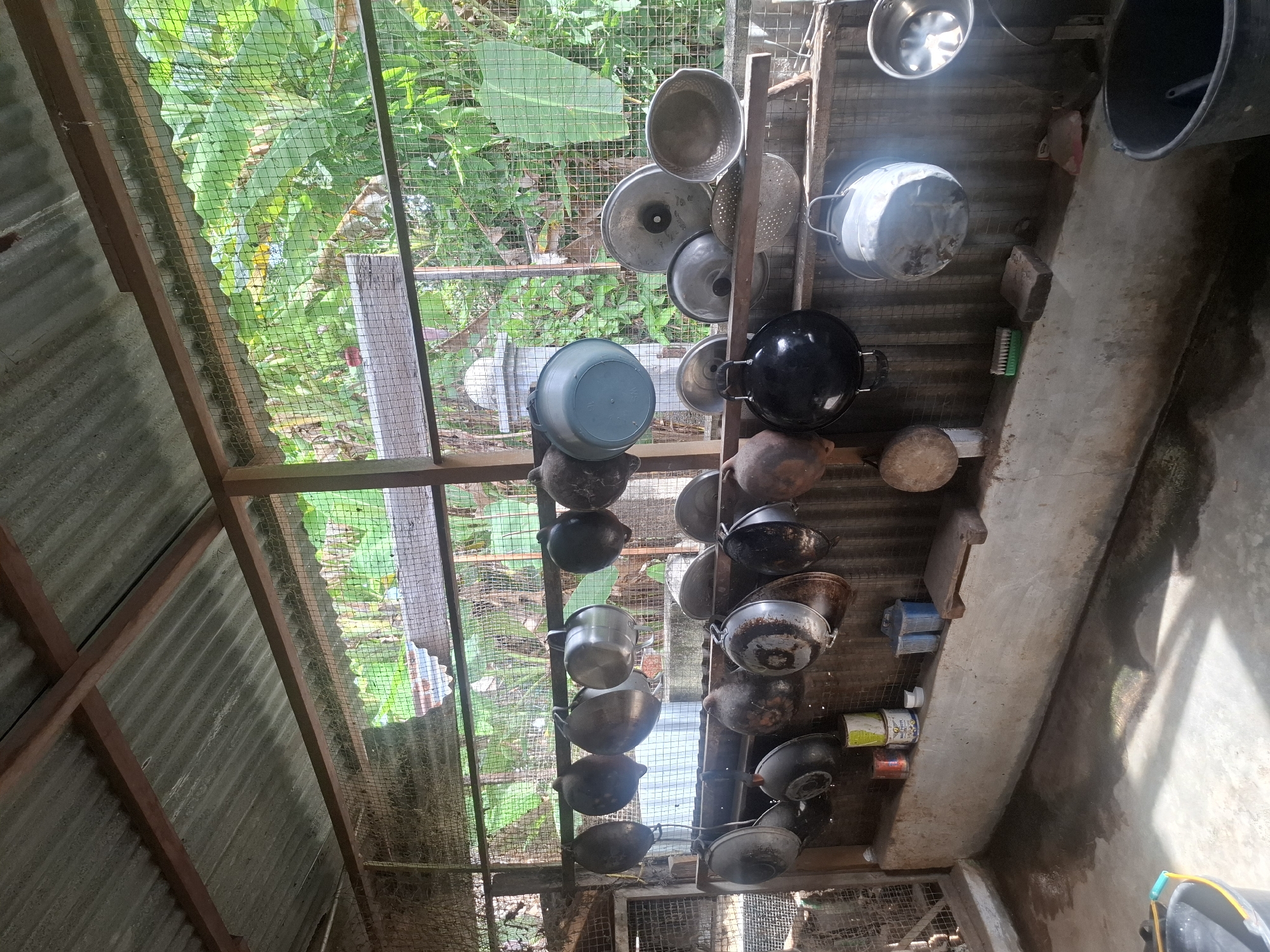 The image depicts an outdoor kitchen area, likely in Jurong, Indonesia, characterized by an open-air structure with a corrugated metal roof supported by wooden beams. The walls are constructed with wire mesh, allowing visibility of lush green vegetation beyond. Various cooking utensils, including pots, pans, and bowls of different sizes and materials such as metal and clay, are hung from racks. A concrete ledge runs along the back wall, upon which several items are placed, including canisters, a blue plastic container, and a small wooden chopping board. The floor is a concrete surface, wet in some areas, with sunlight casting shadows. In the foreground, a dark plastic bucket is visible. The scene appears to be functional and utilitarian, typical of a domestic or small-scale food preparation area in a tropical environment.