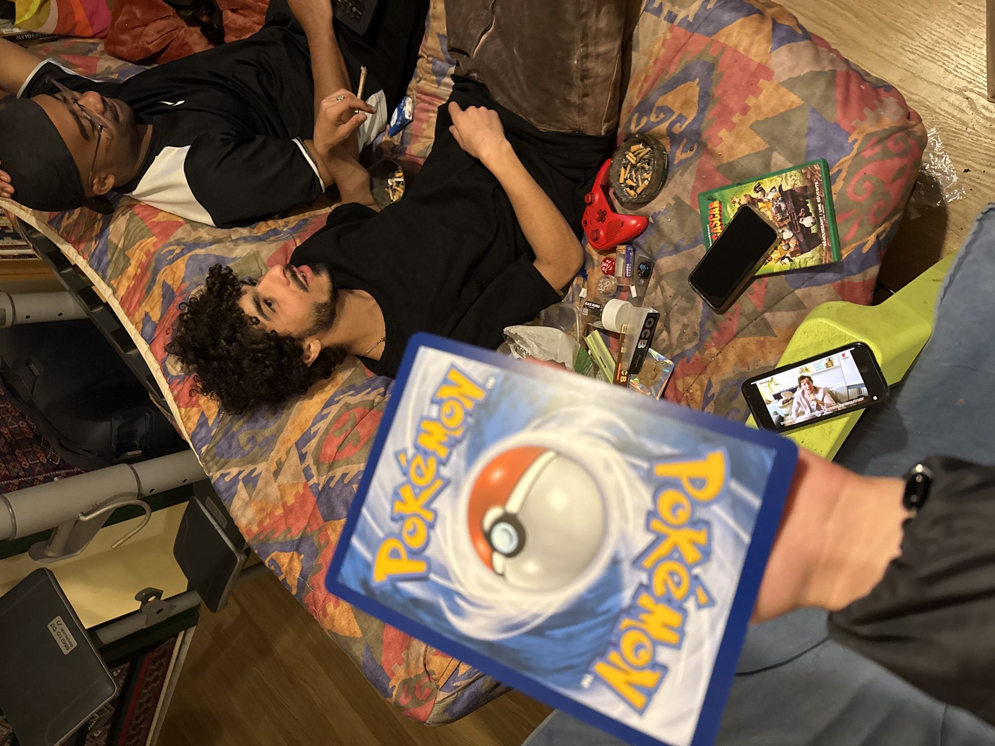 Two young men are lying on a bed with a patterned mattress. One man wears glasses and a black and white shirt, holding a lit joint. The other man has curly hair, a beard, and a black shirt. Beside them on the bed are miscellaneous objects including a red game controller, lighters, rolling papers, an ashtray filled with cigarette butts, a DVD case, and two cell phones. One of the cell phones is on and visible in the foreground, in the hands of a third person who is partially in the frame and who is holding a Pokémon card. The location is not identifiable as Gif-sur-Yvette, France.