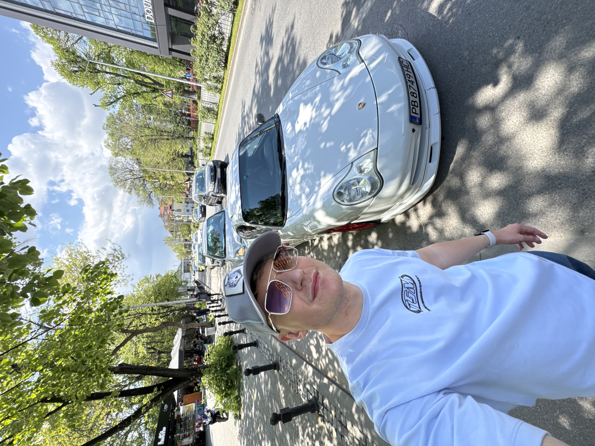 A man takes a selfie on a city street in Plovdiv, Bulgaria, next to a white Porsche. The street is lined with trees, small black bollards, and cafes. The buildings are modern. The man wears a baseball cap, sunglasses, and a white t-shirt. The license plate of the car is "PB 8749 HC". Other vehicles are parked further down the street. The weather is sunny.