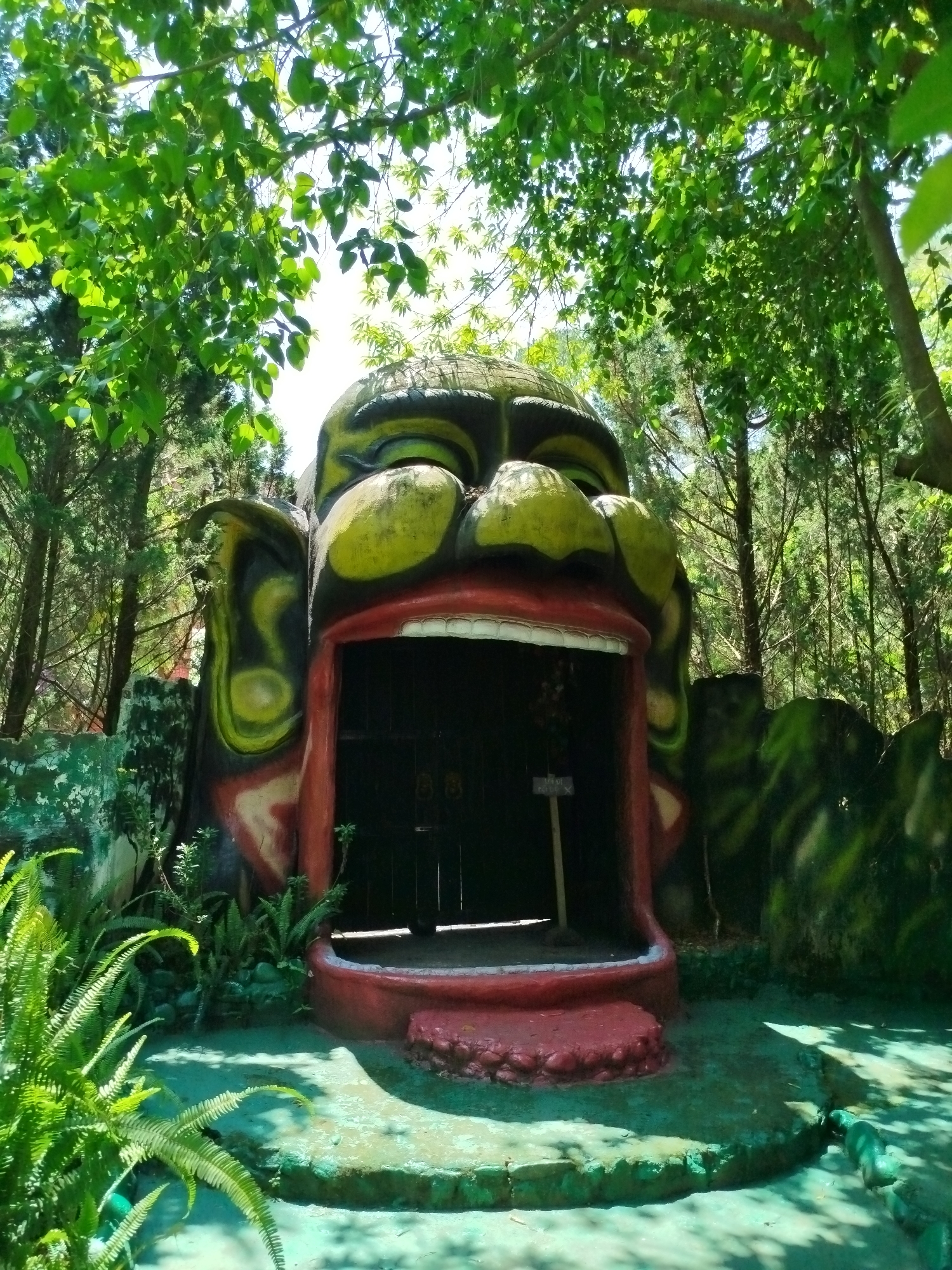 The image shows a large, colorful sculpture of a human head with an open mouth. The sculpture appears to be part of an entrance to a structure in what seems to be a wooded area. The head is painted with green and yellow skin tones, red lips, and black outlines. The mouth serves as a doorway, revealing a dark interior behind a gate. The area surrounding the sculpture is overgrown with various plants and trees. The location is Păng Tiêng 1, Vietnam.