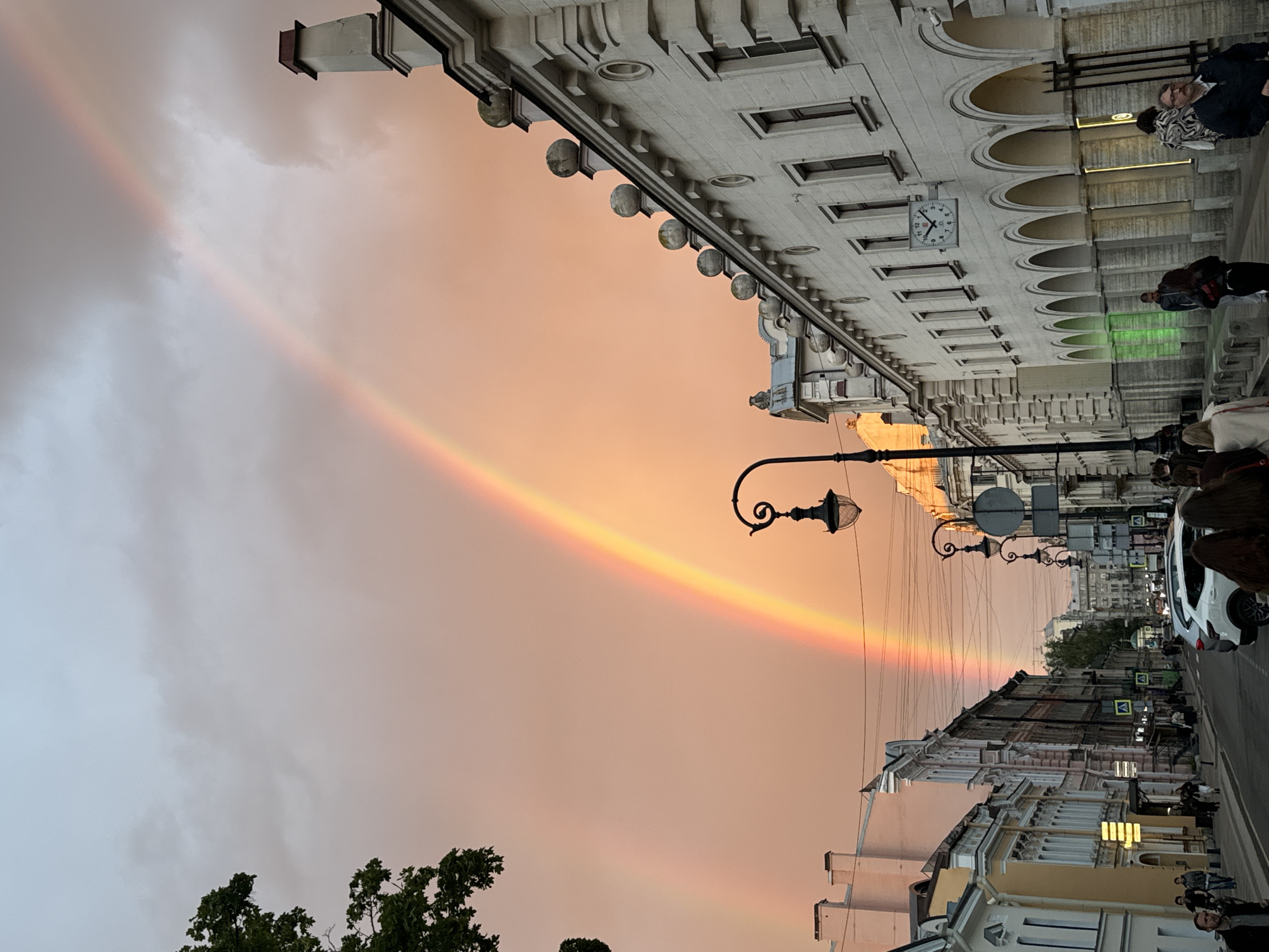 This image captures a street scene in Saint Petersburg, Russia, featuring a prominent double rainbow arching across a sky tinged with the warm hues of sunset or sunrise. The sky transitions from a soft orange to a lighter gray, suggesting a period of transitioning light.
The foreground is dominated by the architecture of historic buildings lining the street. The building on the right displays classical architectural elements, including arched windows and decorative facades. A streetlamp with an ornate design is visible, adding to the vintage feel of the urban environment. Further down the street, more buildings with detailed facades are visible, some appearing older than others. Traffic signals and road signs are present, indicating a functional street. A white car is parked on the side of the road.
Several individuals are present on the street, some walking and others possibly standing near the buildings. Their presence adds a sense of everyday life to the scene. The image is oriented vertically, with the sky and rainbow taking up the upper portion, and the street and buildings filling the lower portion.
The overall mood is one of serene beauty, with the natural phenomenon of the rainbow contrasting with the man-made urban landscape. The soft lighting of the sky and the presence of the rainbow evoke a sense of wonder and tranquility. No specific text is clearly legible in the image.