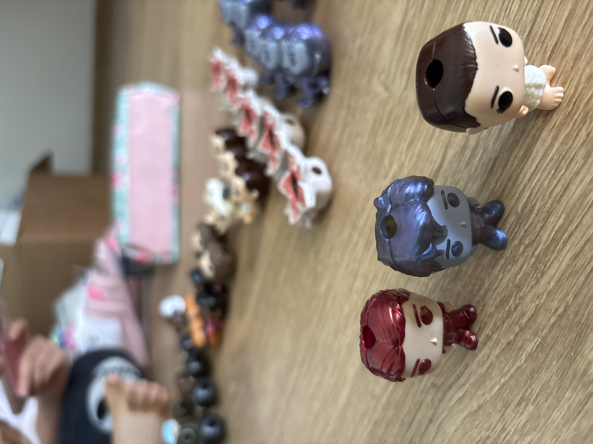 A photograph shows a collection of toys or figurines, some arranged in a line or row on a light-colored wooden surface. At least three figurines are oriented facing up in the frame, these are close to the foreground, while others are arranged to the left and further in the background. The toys or figurines include human figures and animal shapes, possibly dragons. The human figures on the right side have prominent round heads and small bodies, and include details like dark hair, skin tones, and stylized features. Some figures are metallic.
In the background there are boxes and other materials, and a person's hands can be seen. The person is wearing a dark t-shirt and white sleeves.
The exact location in the city of Fulda, Germany cannot be determined from the image content alone. The environment appears to be indoors.