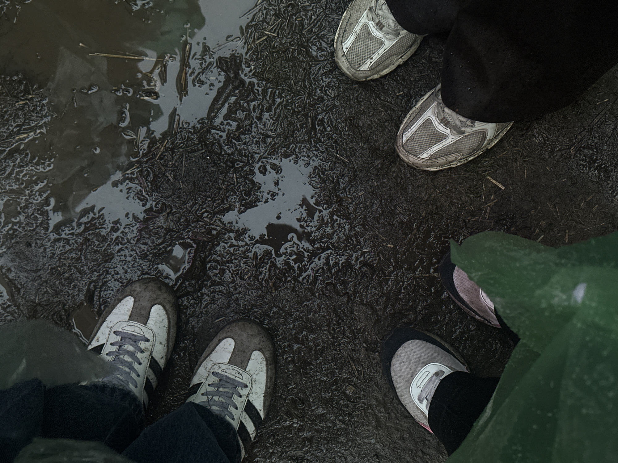 The image shows the feet of at least four individuals standing on muddy ground with puddles of water. They are wearing various athletic shoes, some of which are dirty. The ground appears to be a mix of mud, water, and organic debris like straw or grass. One individual is wearing a green garment, possibly rain gear. The lighting is dim and overcast. No specific landmarks or details are visible to indicate a location within Ivano-Frankivsk, Ukraine, or any other specific city. The scene depicts an outdoor environment that is wet and muddy, suggesting recent rain or proximity to water.