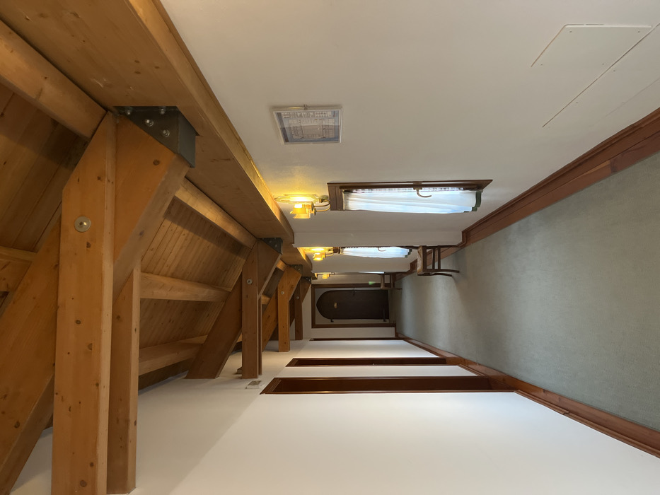 The image shows a hallway with exposed wooden beams, gray metal brackets, and fasteners on the left side. The beams are connected to the ceiling. The right side has a gray wall with a wooden trim. There are several windows with white curtains along the ceiling. Further down the hall, there is a dark wooden door. The walls are painted white. There are wooden accents on the floor.