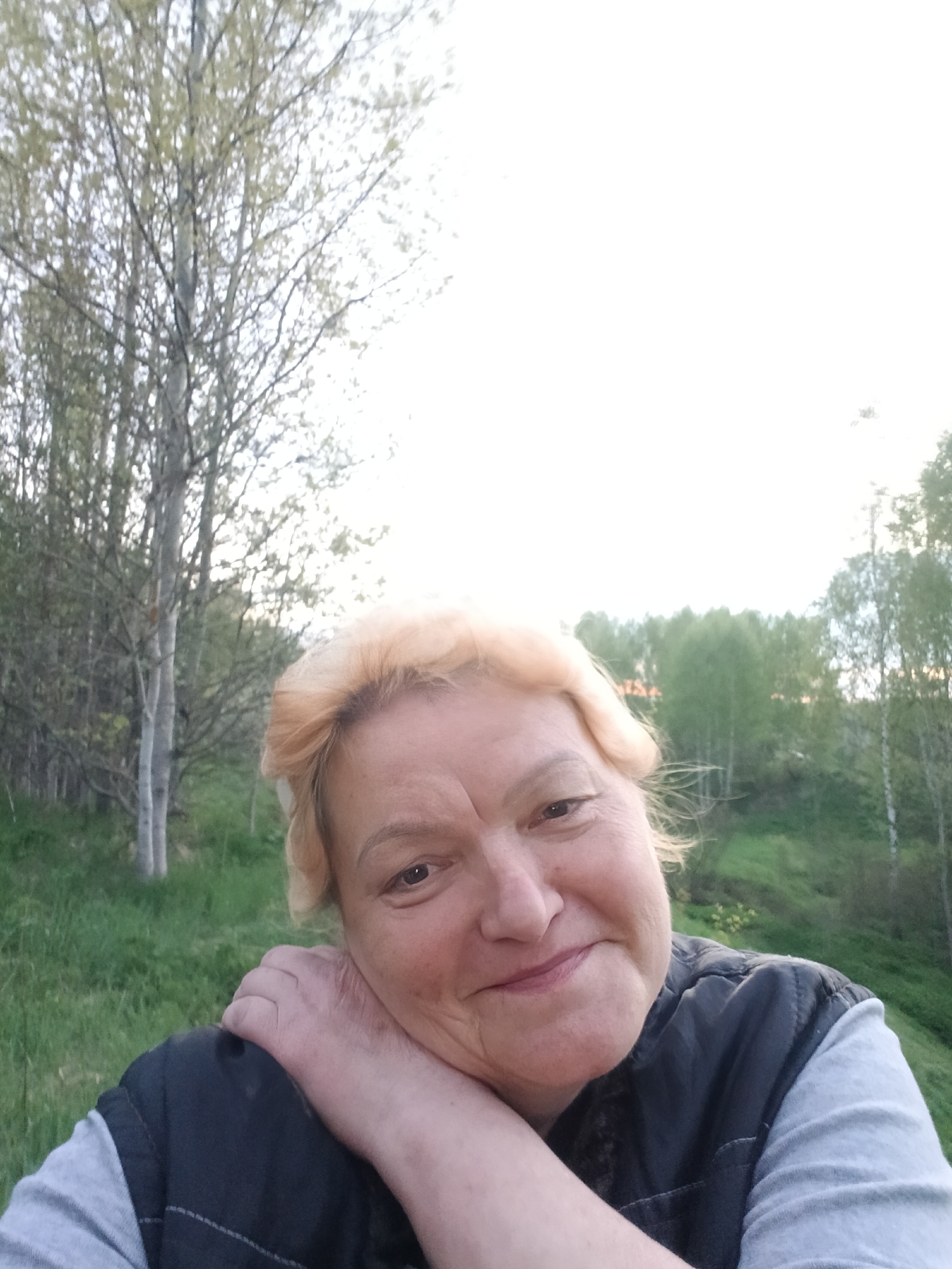 A middle-aged woman with short, light blonde hair and fair skin smiles at the camera, resting her face on her hand. She is wearing a gray t-shirt and a dark vest. Behind her is a grassy hillside dotted with trees in various shades of green. In the background, more trees line the horizon. A large birch tree is visible to the left of the woman. The sky is bright and slightly overcast. The photograph appears to have been taken during the day in Красный Маяк, Russia.