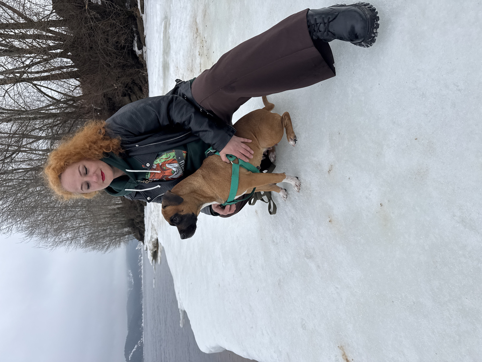 A woman with red, curly hair and wearing a black leather jacket, green hoodie with graphic, and brown pants stands on a snowy surface with a tan dog with a black face and white paws. The dog is wearing a green harness. The woman's left hand is on the dog's back. The location appears to be a riverbank covered in snow, with trees visible in the background and a body of water extending into the distance under a cloudy sky. It is likely on the city of Chaikovsky, Russia.