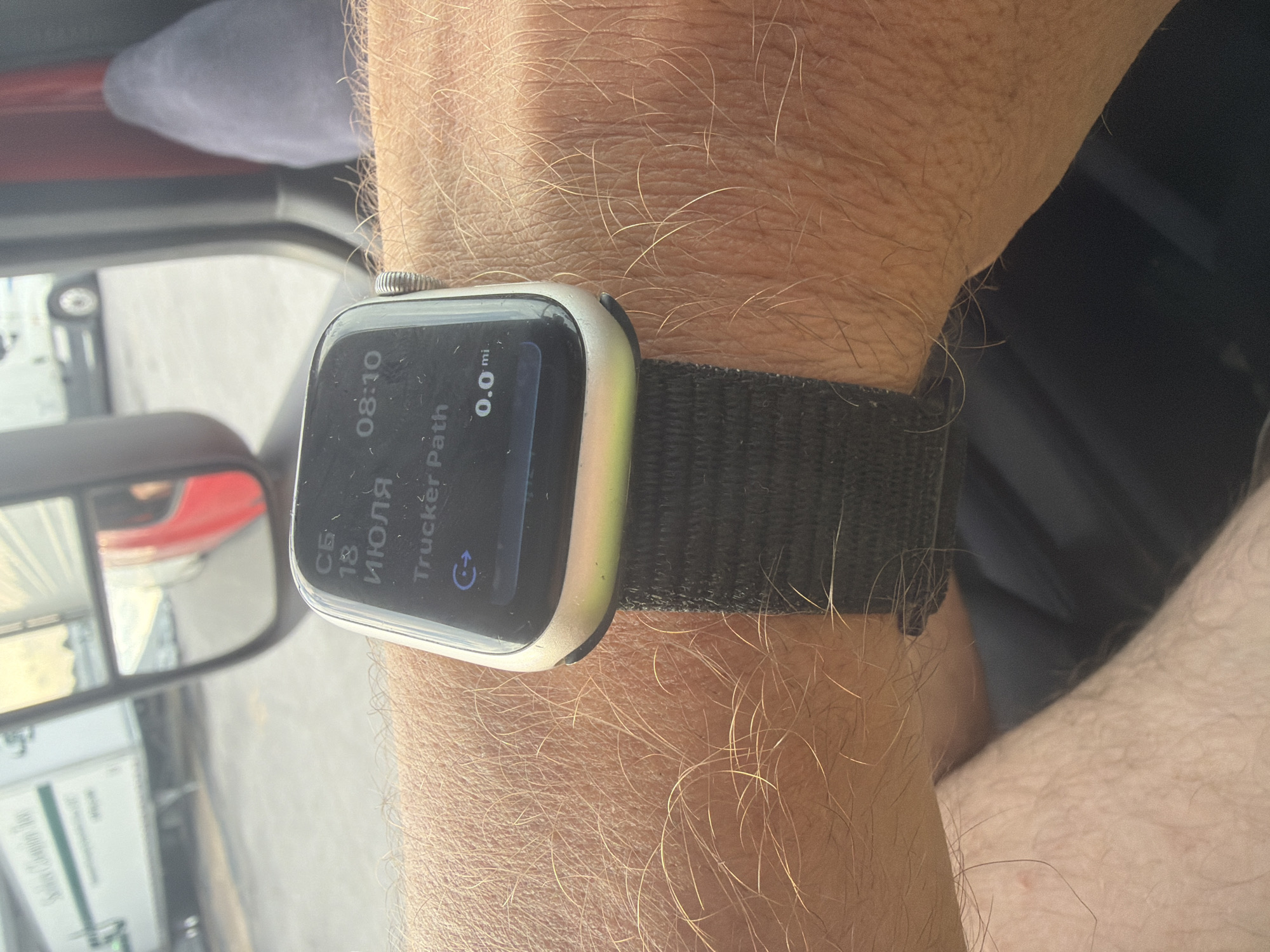The image shows a close-up of a person's arm wearing an Apple Watch. The watch displays the time as 08:10 and the date as July 18th. It also shows "Trucker Path" with a distance of 0.0 miles. In the background, a semi-truck's side mirror reflects a red semi-truck cab. The environment appears to be outdoors, possibly in a truck stop or parking lot, with other trucks visible in the background, suggesting a transportation hub. The location is the city of Chattanooga, United States of America.