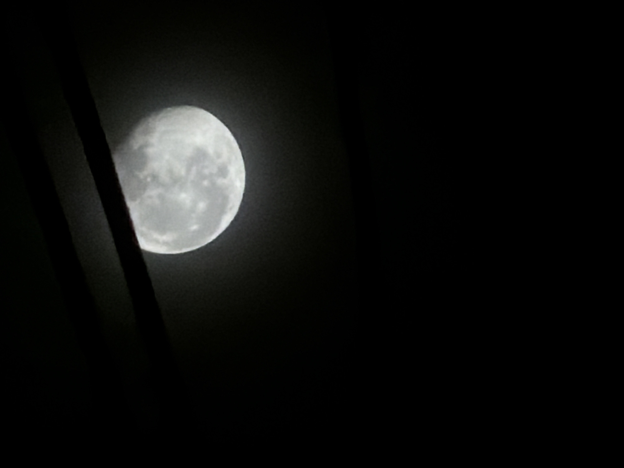 The image displays a nocturnal scene featuring a large, bright Moon against a dark sky. The Moon, appearing to be in a gibbous phase, is positioned in the upper-left portion of the frame, partially obscured by dark, silhouetted objects. Its surface details, including craters and darker maria, are discernible, indicating a clear atmospheric view.
Two prominent, dark, linear structures run diagonally from the top-left towards the bottom-center of the image, partially covering the Moon and the surrounding dark expanse. These objects are in deep shadow, rendering them indistinct, but they appear solid, possibly representing parts of a building, poles, or thick tree limbs, suggesting the photograph was taken from an obstructed vantage point. The rest of the background is an undifferentiated deep black, typical of a moonlit night sky without other visible celestial bodies.
The scene depicts a quiet night. There are no people or discernible activities taking place, focusing solely on the celestial body and its immediate foreground obstructions. The time of day is clearly night, with clear weather conditions allowing for the Moon's visibility. There are no specific location cues within the image itself to identify it as Bangkok, Thailand, and no visible text is present.