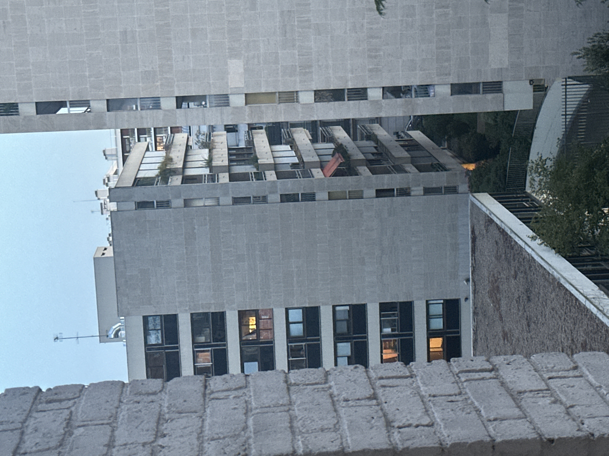 The image shows a view of apartment buildings in an urban setting. The buildings are tall and made of concrete, with many windows and balconies. Some of the windows are lit, suggesting that people are inside. In the foreground, a brick wall is visible, which appears to be part of another building or structure. The sky is overcast, and the overall lighting suggests it is either early morning or late evening. The scene is devoid of any discernible human activity or prominent objects.