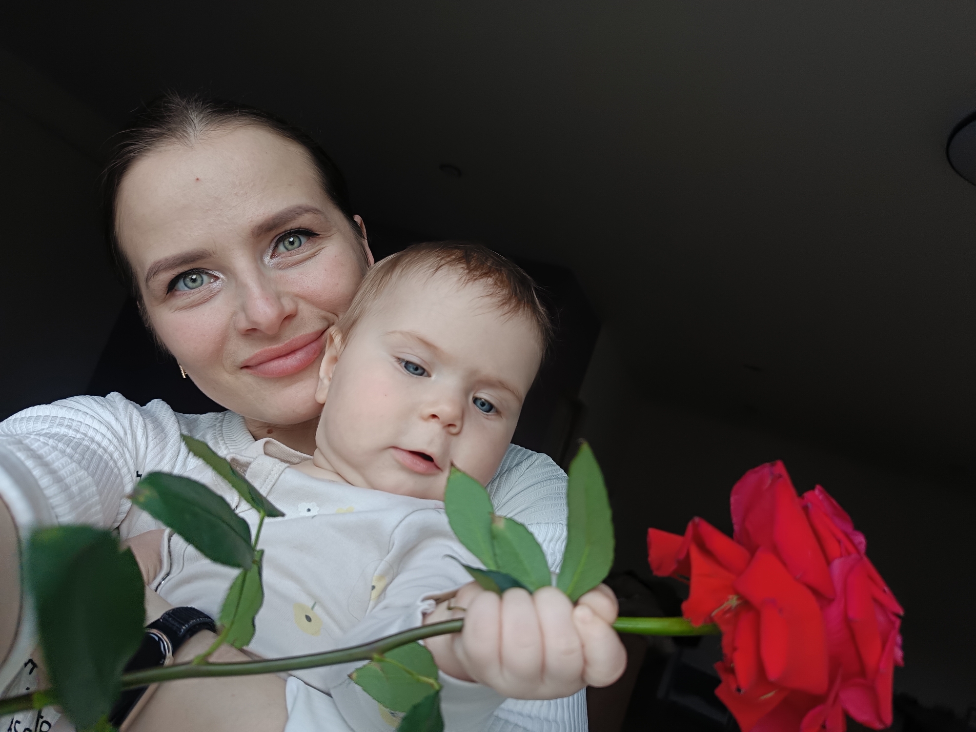 A heartwarming, close-up indoor portrait captures a mother holding her baby. The mother, with dark hair pulled back and striking blue-green eyes, smiles softly towards the camera. She is wearing a white, ribbed long-sleeved top, and a black wristband or watch is visible on her left wrist. The baby, nestled against the mother, has light, possibly reddish-blonde hair and bright blue eyes, and wears a light-colored onesie with subtle yellow patterns.
The baby is actively engaged, holding the green stem of a vibrant red rose with both hands, seemingly fascinated by it. The rose head, full and slightly textured, extends towards the right side of the frame, with several prominent green leaves along its stem. The background is largely dark and out of focus, creating an intimate ambiance, indicative of a low-light indoor setting. The image conveys a tender moment of affection and curiosity between mother and child, with the rose acting as a central point of interest. There is no visible text in the media. The provided location context is Rusalevka, Russia.