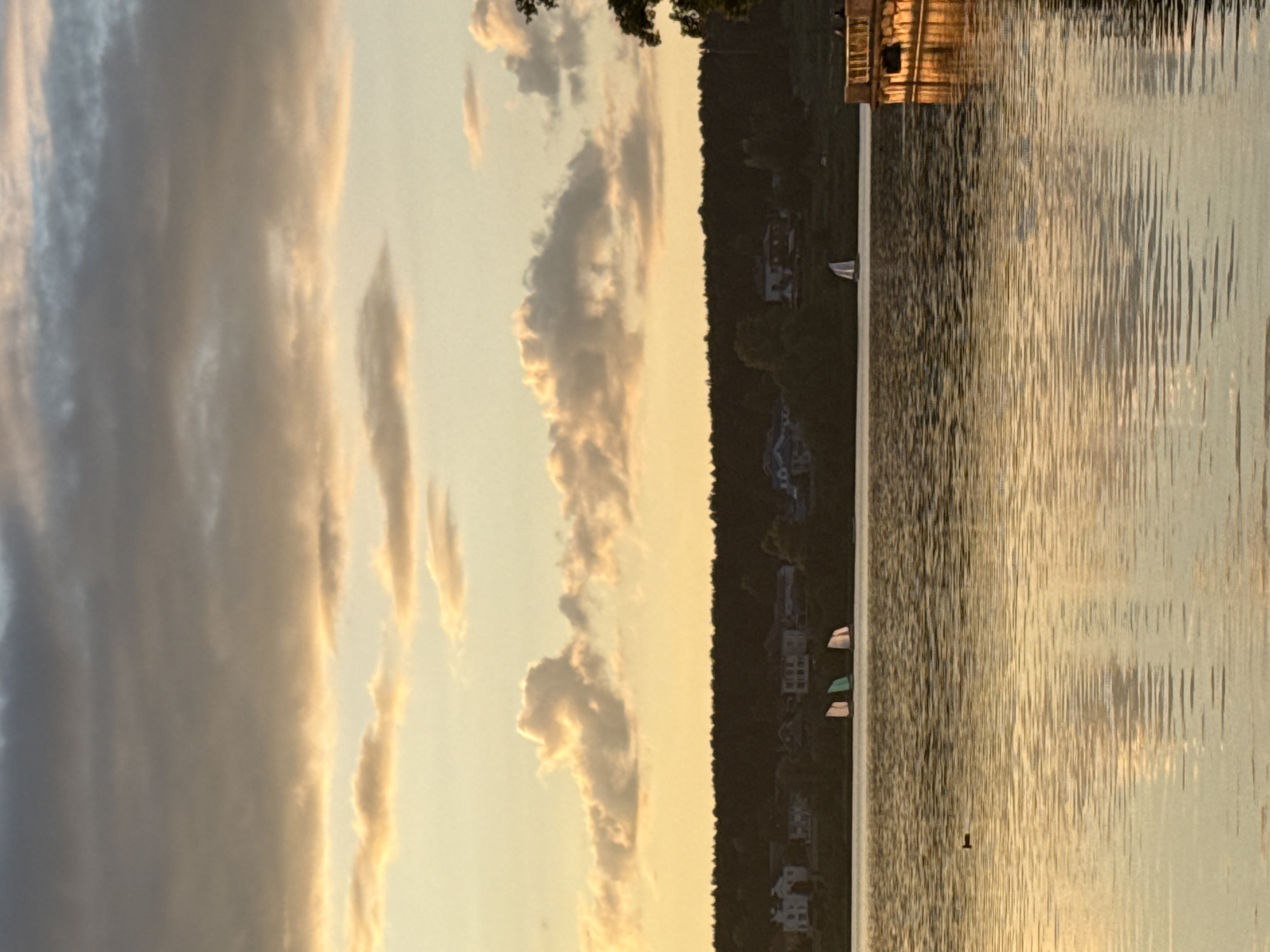 The image, set at golden hour over the Votkinsk Reservoir in Russia, depicts a tranquil horizontal landscape dominated by a sunset sky and calm water. The upper half features a clear sky with scattered, elongated clouds illuminated from below by warm, golden light, creating a serene atmosphere. The lower half is filled with the reflective surface of the water, shimmering with the golden hues of the setting sun and showing subtle ripples. A dark, forested shoreline stretches across the middle ground, separating the sky from the water. Amidst the dense trees, several distant houses or buildings are visible. On the water near the distant shore, a few small boats, including a white sailboat, are present. A wooden dock or pier extends from the right into the water, catching the warm light. A small, dark object floats on the water closer to the viewer. The scene suggests a peaceful evening, with no explicit human activity or visible text.