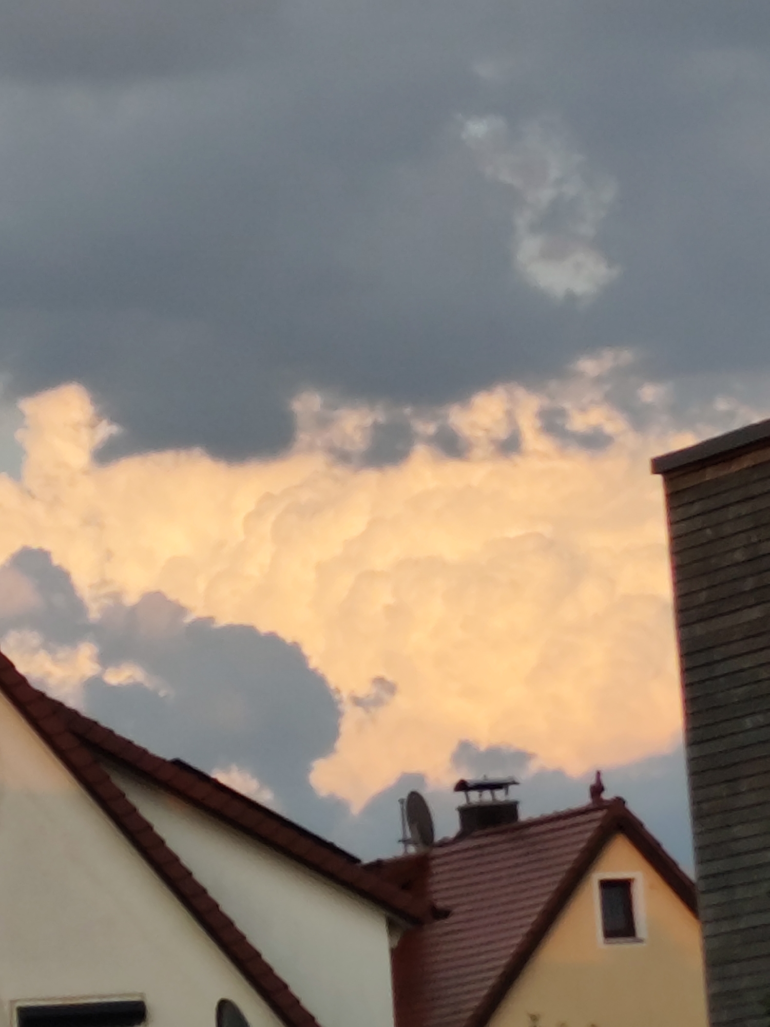 The image shows a cloudy sky over rooftops in Dillingen an der Donau, Germany. The clouds are a mix of dark gray and bright white, with the white clouds illuminated by what appears to be sunset light. In the foreground, parts of two houses are visible, one with a white facade and a red-tiled roof, and another with a yellow facade and a red-tiled roof. A chimney with a satellite dish is on the yellow house. The sky occupies the majority of the frame.