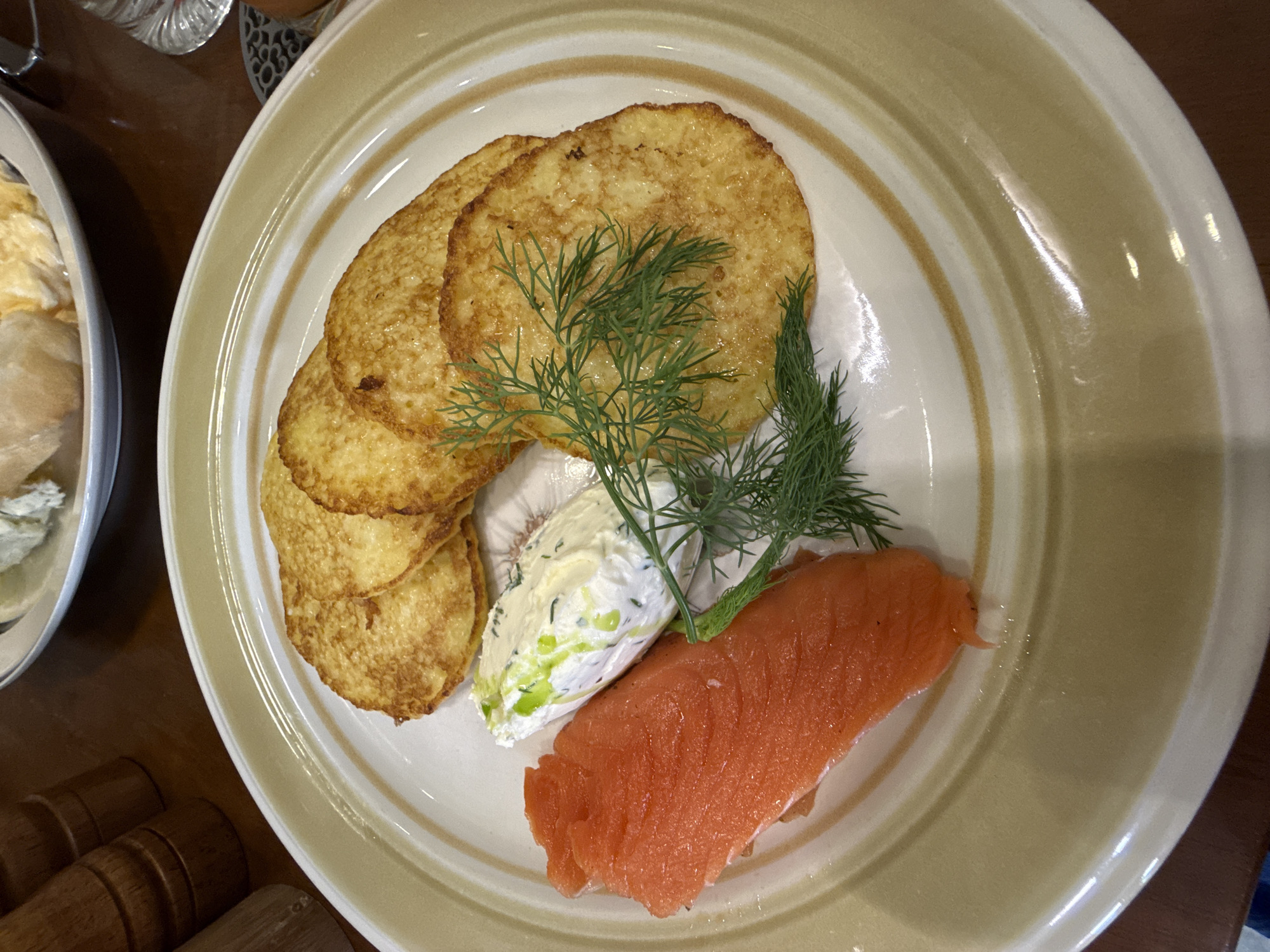 This close-up, overhead shot displays a beautifully plated meal, likely being enjoyed indoors in Nha Trang, Vietnam. The central focus is a white ceramic plate adorned with subtle light brown concentric rings. On the plate, three golden-brown, round pancakes or blinis are stacked on the left side. A vibrant portion of smoked salmon, rich orange in color, rests on the lower right. Between these components lies a dollop of creamy white sauce, which appears to be a herb-infused sour cream or cream cheese, featuring visible specks of green herbs. Fresh sprigs of green dill are elegantly draped across the blinis and the creamy sauce, serving as a garnish.
The food is meticulously arranged, suggesting a carefully prepared dish from a restaurant or a thoughtful home-cooked meal. To the left of the main plate, a partial view of another white dish or bowl reveals a soft, light-colored food item, possibly scrambled eggs or another dairy-based dish. The background subtly shows elements of a dining setting, including a wooden table surface and hints of other items like a clear glass and wooden kitchenware, possibly salt and pepper grinders, in the lower left corner. The lighting is soft and even, illuminating the textures and colors of the food without harsh shadows. No individuals are visible, and there is no discernible text within the image. The overall impression is one of a fresh, appealing, and well-composed meal.