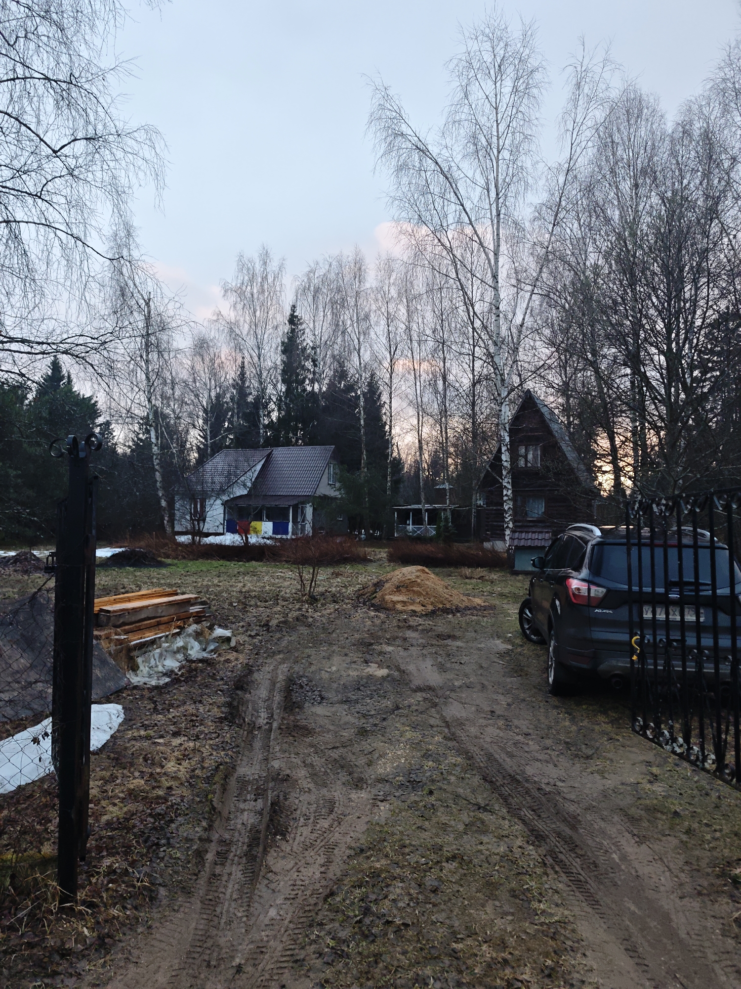 The image depicts an exterior scene in Свистуха (кузяевский С/о), Russia. The ground is a muddy track with clear tire tracks. To the left, there is a pile of wooden planks covered partially by a white sheet or tarp. A rustic metal fence and gate are prominent in the foreground on both sides.
In the background, two houses are visible. The first is a white building with a brown roof, and colorful material hanging from the porch. The second is a dark wooden structure with a peaked roof.
A dark-colored SUV is parked beside the fence on the right. The license plate is partially visible, but not decipherable.
The scene is surrounded by bare trees, suggesting it is either late autumn, winter, or early spring. The sky is overcast with hints of pink towards the horizon.