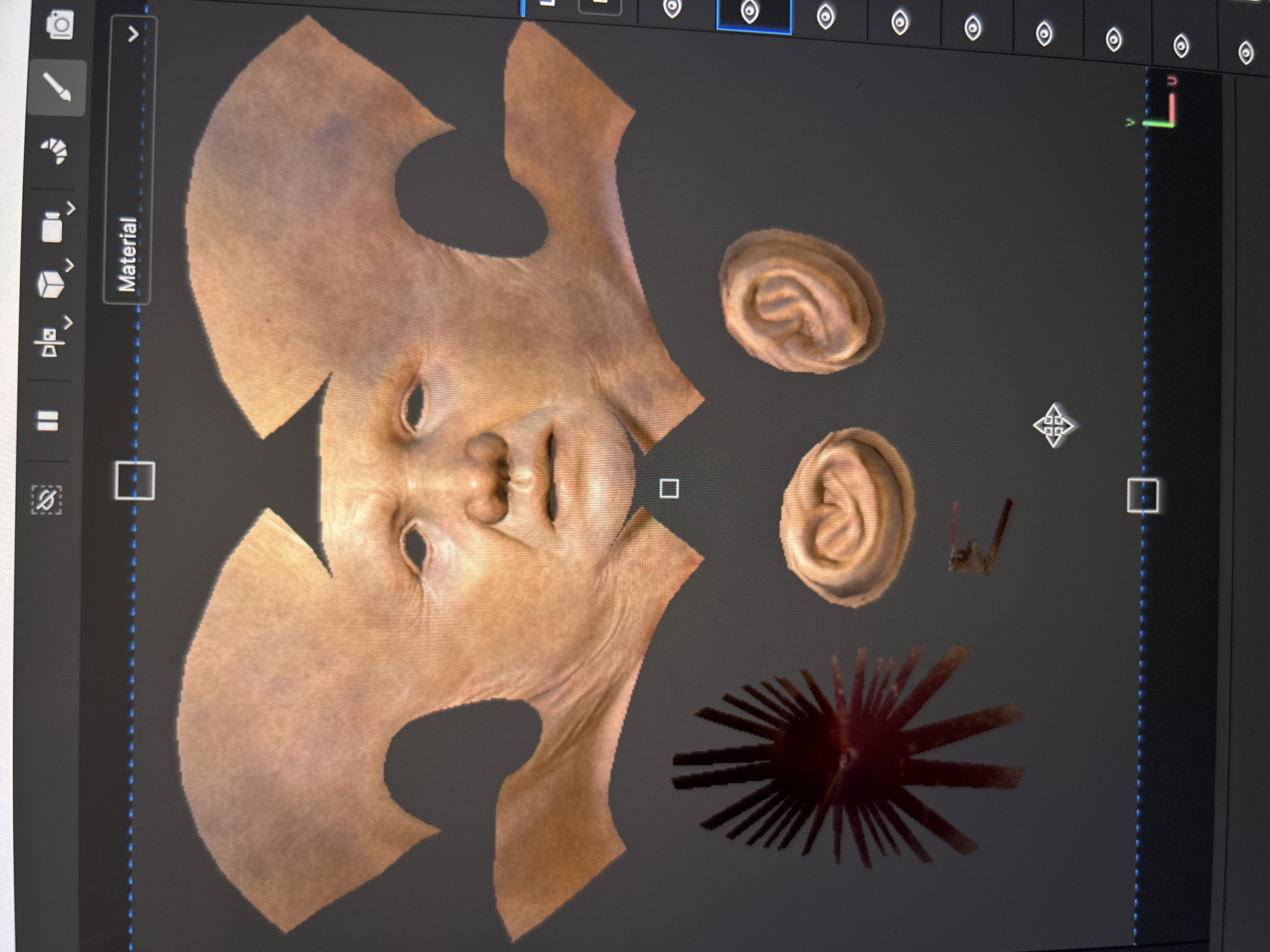 The image displays a computer screen showing a 3D modeling or texturing software interface. The main subject is an unwrapped 3D texture map of a human-like face, rendered with realistic skin tones, pores, and wrinkles in a light brown/tan hue. A subtle bruise or discoloration is visible on the forehead area of the unrolled texture.
Alongside the face, there are two separate 3D models of human ears, also textured with skin-like details. Towards the bottom right of the screen, a distinct red, spiky 3D object, possibly an accessory or part of a creature, is visible.
The scene implies an activity of 3D modeling, texturing, or UV mapping. The software environment features a dark grey background. Various user interface elements are visible, including a vertical toolbar on the left with icons such as a camera, a brush, and a cube. A prominent vertical tab labeled "Material" is present on this toolbar. The top toolbar displays several eye icons, likely for toggling visibility. A 3D coordinate system (X, Y, Z axes) is situated in the top right corner, and a square selection tool is visible in the center of the display. A vertical dotted blue line runs through the screen, possibly indicating a selection boundary or guide. The lighting on the 3D models appears neutral and even, designed to showcase the textures clearly. The photo was taken in Cape Coral, United States of America.