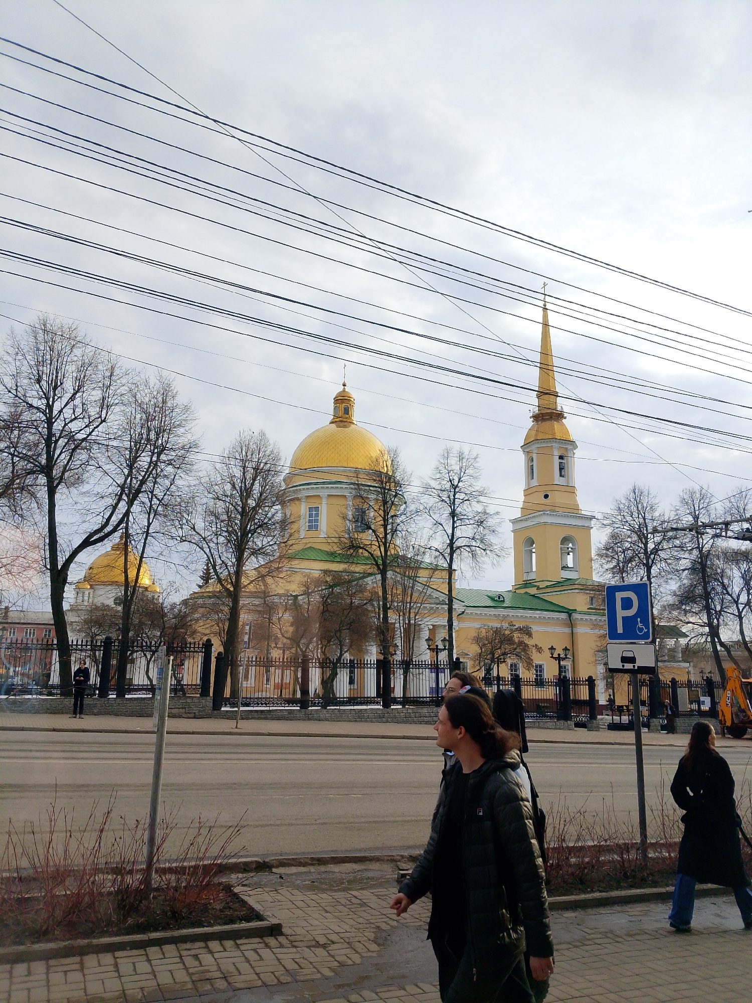 The image shows a street scene in Izhevsk, Russia, dominated by the Alexander Nevsky Cathedral in the background. The cathedral is a large, yellow building with gold domes and a tall, slender bell tower topped with a spire. Power lines stretch across the sky in the upper portion of the frame. In the foreground, several people are walking along the sidewalk. A parking sign for the disabled is visible near the curb. On the far side of the street, a construction vehicle is parked near the church. Bare trees are interspersed throughout the scene, suggesting a cool season. The sky is overcast.