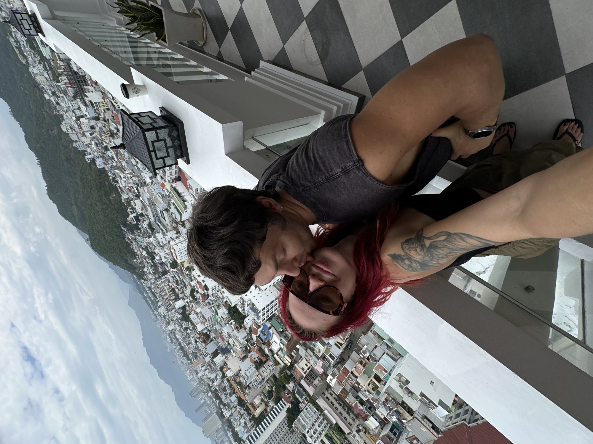 A couple is on a balcony overlooking the city of Nha Trang, Vietnam. The man is kissing the woman on the cheek. The woman has red hair and is wearing sunglasses and a black top. The man is wearing a dark t-shirt. The balcony has a railing and a checkered tile floor. In the background, the city stretches out to the mountains. There is a planter with a snake plant and two decorative lanterns on the balcony. The sky is cloudy.