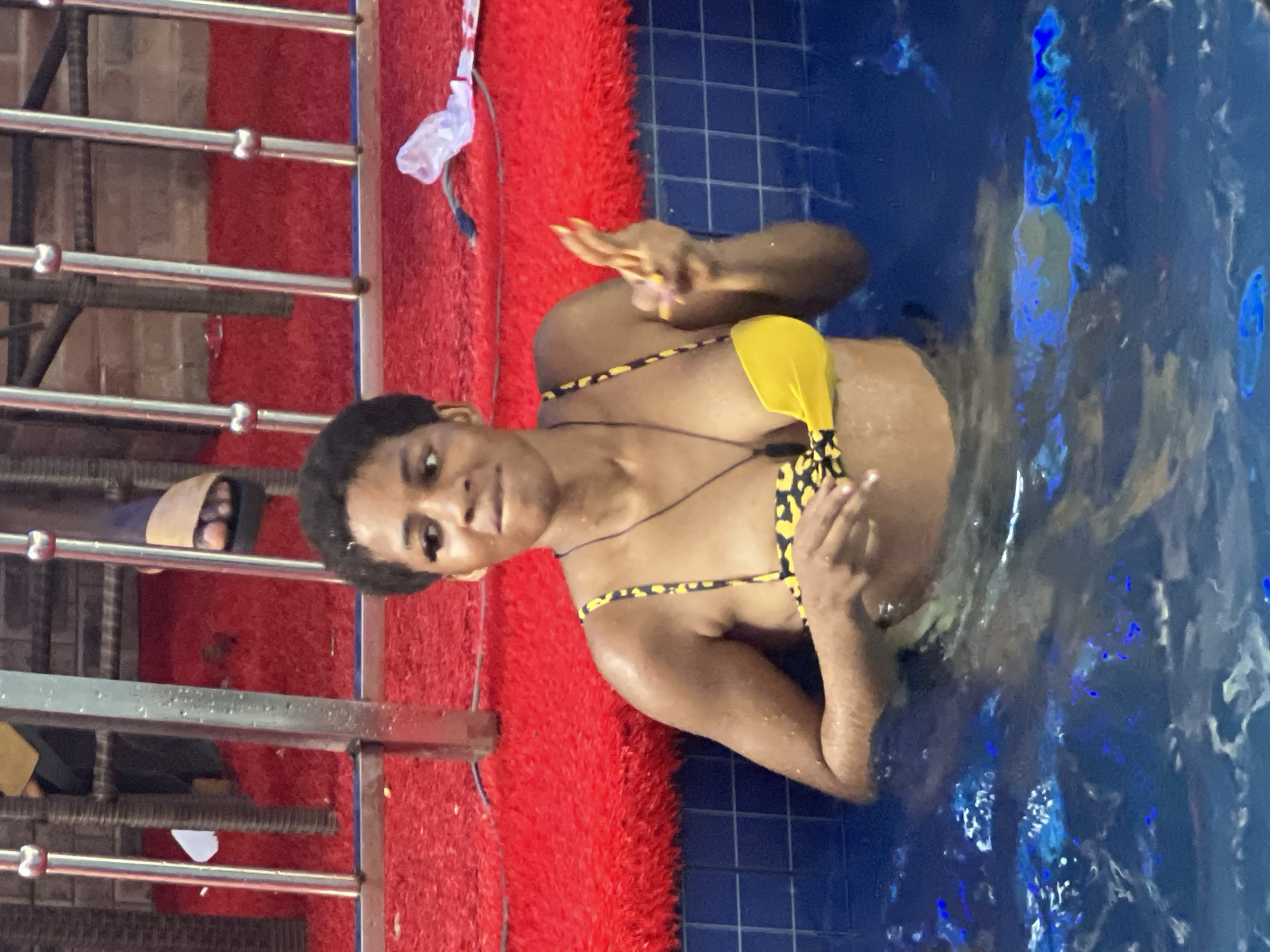 A young Black woman is in a swimming pool, wearing a yellow and black animal print bikini. She is partially submerged in the water and looking towards the camera with a neutral expression. Her right hand is raised with her fingers spread, and her left hand is near her chest. The pool water is a deep blue with some blue lighting effects visible underwater. The edge of the pool is lined with blue tiles. To the left of the pool, there is a red, fuzzy mat, and behind that, a metal railing. The railing is situated in front of a brick wall. The scene appears to be outdoors, possibly during the daytime, though no clear time of day or weather cues are visible beyond the water itself. The location context is Obosi, Nigeria. There is no visible text in the image.