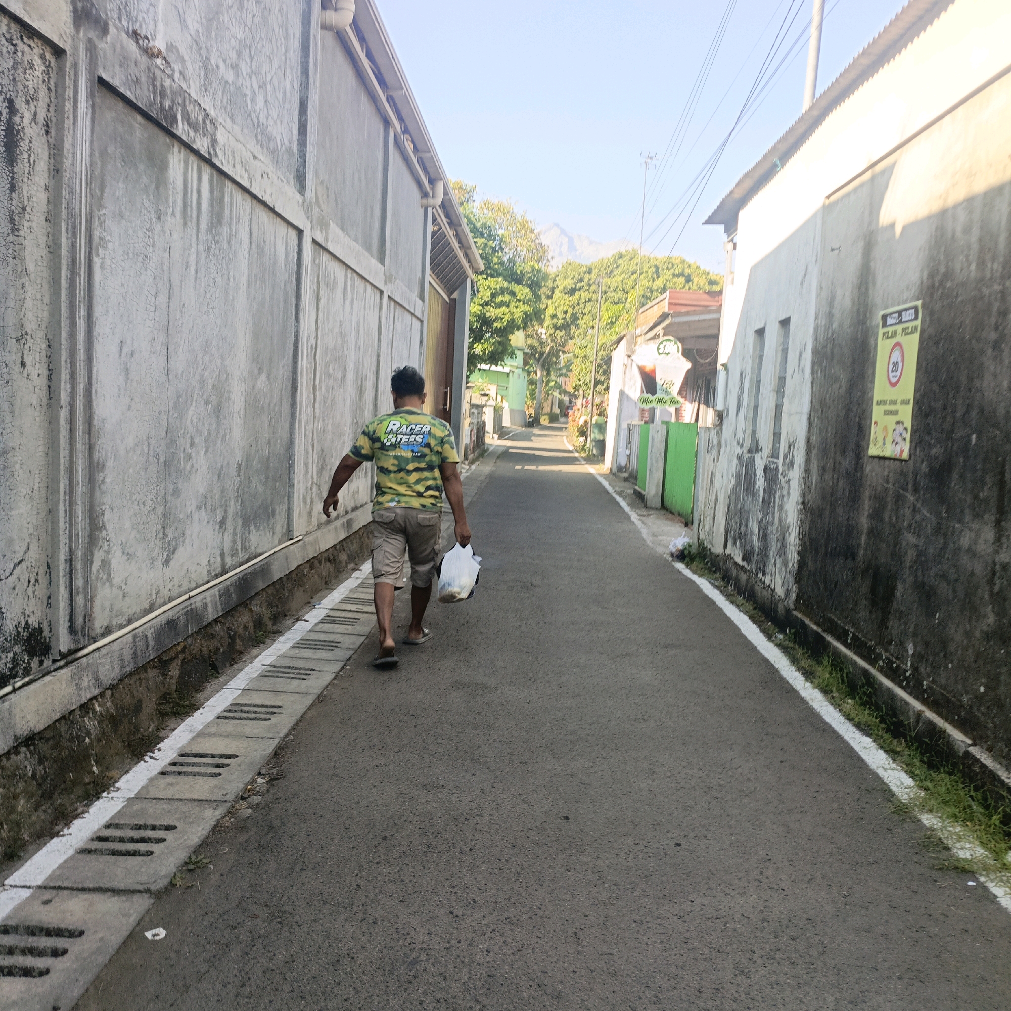 A man walks down a narrow, paved street lined with buildings and trees in Salatiga, Indonesia. The man, seen from behind, wears a camouflage-patterned t-shirt with the words "Racer Tees" and "Team" visible, and khaki shorts. He carries a white plastic bag in his right hand. The street has a concrete drainage channel with grates along the left side, bordered by a white line. The buildings on either side are made of concrete or stucco, some appearing aged and weathered, with a corrugated metal roof visible on the right. Green planters and fencing line the right side of the street further down. In the distance, trees and a mountain can be seen under a clear blue sky. A yellow sign with black text and a red circle with the number 20 is affixed to the wall of the building on the right.