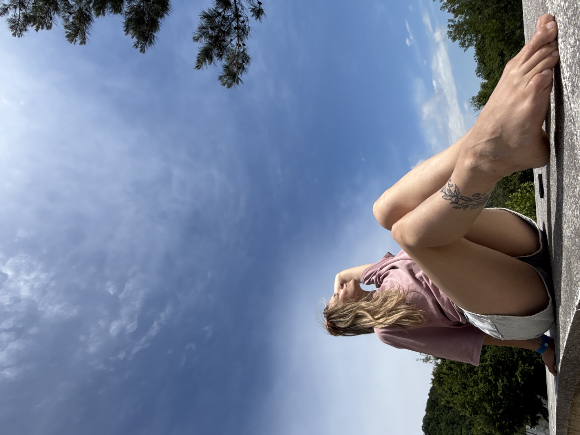 A woman with blonde hair sits on a stone ledge outdoors, looking up at the sky. She is wearing a pink t-shirt and denim shorts. Her right arm is bent behind her head, and her legs are crossed. A tattoo of a leafy vine is visible on her left ankle. The background consists of a bright blue sky with scattered clouds, green trees, and a hint of mountains.