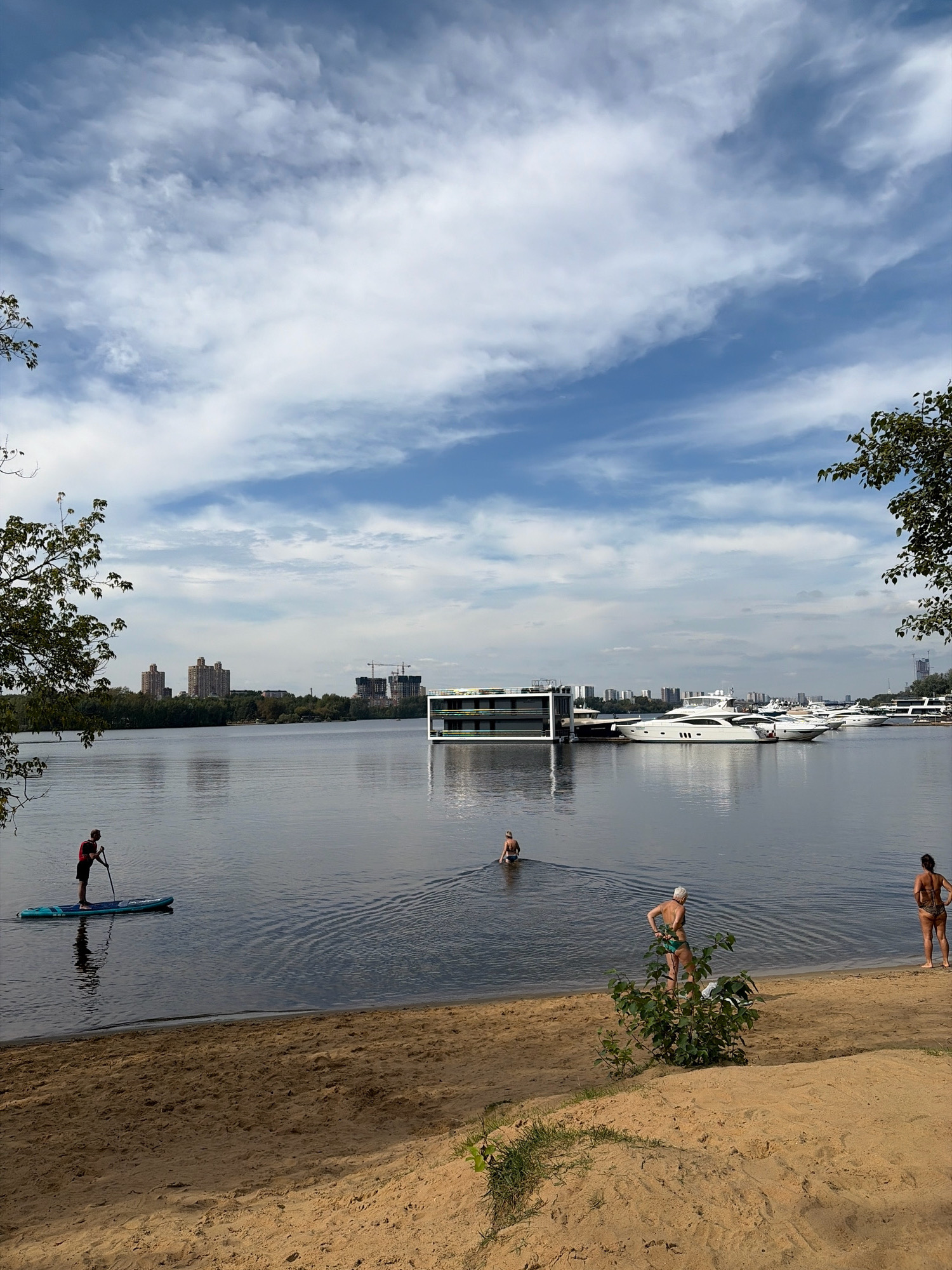 This is an outdoor, landscape, daytime photograph taken from a sandy beach on a body of water, likely a river or reservoir, in Khimki, Russia. The sky is a bright blue with scattered white clouds. In the middle ground, across the water, there is a city skyline with several tall buildings and a few construction cranes visible. Further to the right, a modern, multi-story structure with a white facade and large windows is situated on the water, possibly a floating building or a houseboat. A row of white yachts is docked near this structure.
On the water in the foreground, a person is paddleboarding from left to right, casting a reflection on the calm surface. Further out on the water, two other individuals are visible; one is wading, and another is swimming. On the sandy beach in the right foreground, two people are standing, with one facing away from the camera and the other facing toward the water. A small green bush grows in the sand between them. The sandy beach occupies the bottom half of the frame, with some sparse green vegetation near the bottom right corner.