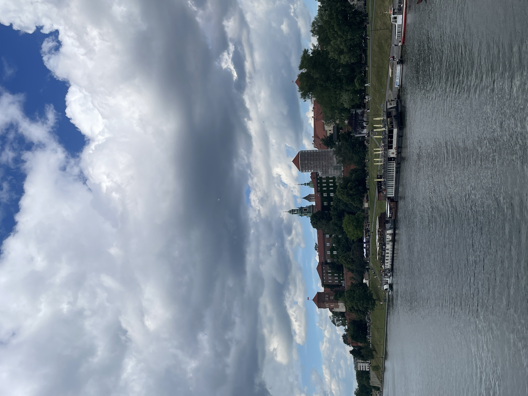 This image shows Wawel Castle and Cathedral situated on a hill overlooking the Vistula River in Krakow, Poland. The castle complex features red-tiled roofs, brick towers, and a prominent green-domed tower. The riverfront is lined with trees and a promenade where several boats are docked, including a large, multi-decked tour boat with yellow umbrellas on its upper deck. People are visible walking along the promenade. The sky is partly cloudy with dramatic, dark storm clouds gathering, interspersed with patches of blue sky and sunlight. The overall scene depicts a historic European cityscape under a dynamic and moody sky.