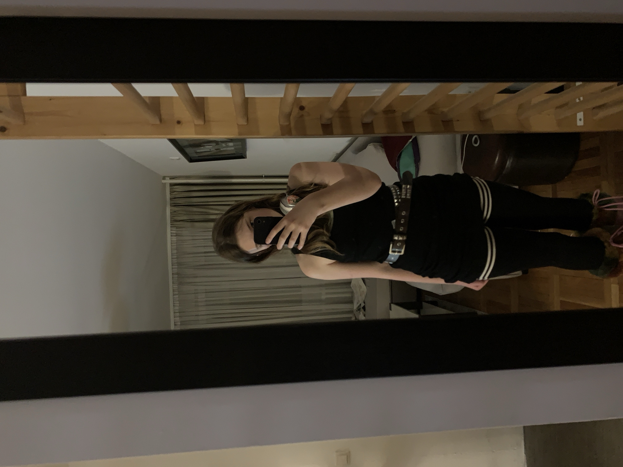 A young woman is taking a mirror selfie in a room. She is wearing a black tank top, black leggings with white stripes, and a studded belt. The room contains a wooden bed frame, a brown drum, and a framed picture on the wall. The location is likely Vozdovac Urban Municipality, Serbia, but this cannot be definitively confirmed from the image.