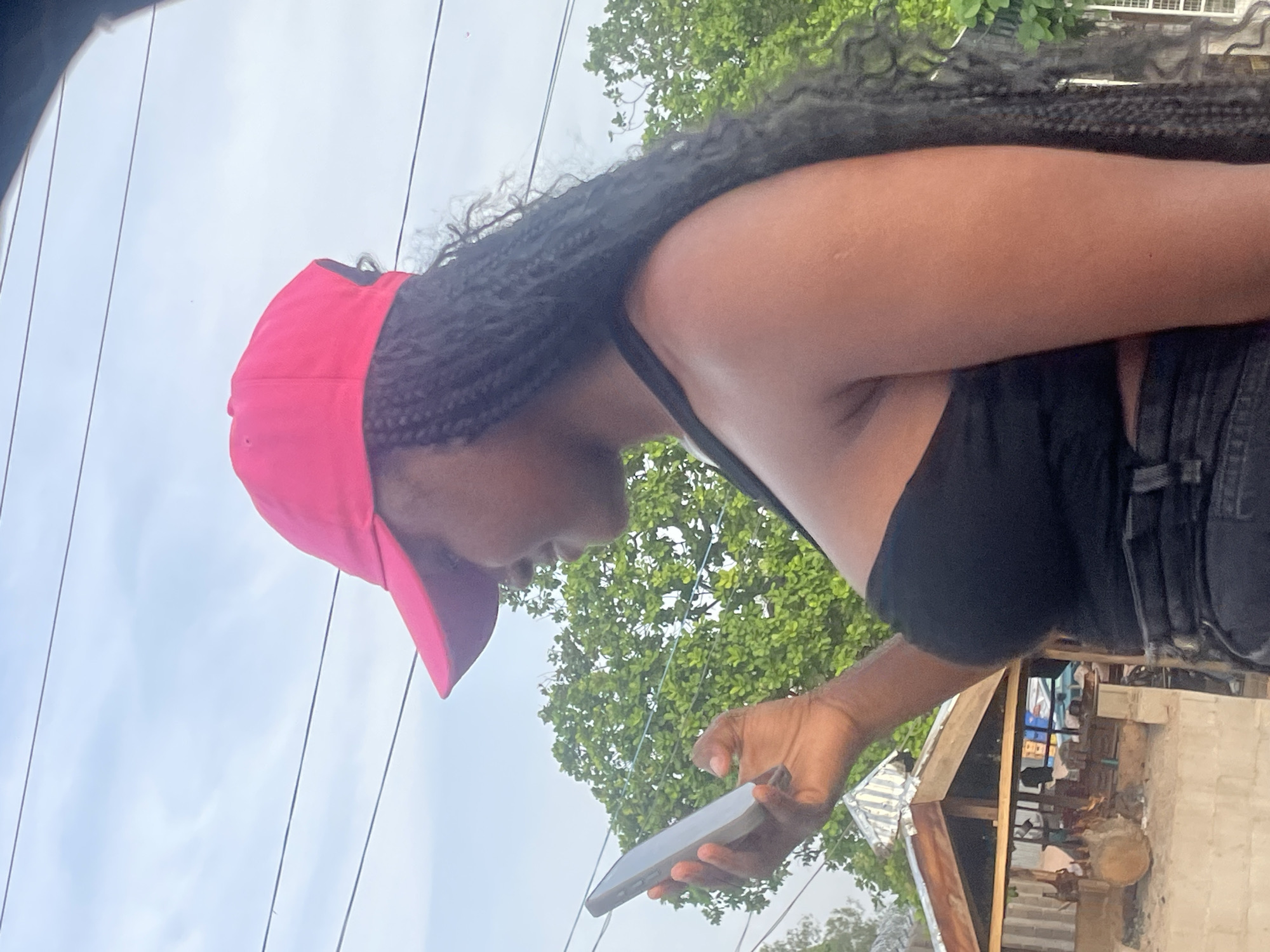 A young Black woman with long, braided hair wears a bright pink baseball cap and a black strapless top. She is looking down at a silver smartphone held in her left hand, her fingers appearing to interact with the screen. Her right arm is bent at the elbow, her hand resting on her lower back. She is positioned in profile, facing left. In the background, lush green trees dominate the middle ground. Below the trees, a modest, open-air structure with a corrugated metal roof is visible, with logs and what appears to be tools or equipment situated in front of it. The scene is set against a pale, overcast sky with thin, horizontal power lines crisscrossing the upper portion of the frame. The environment suggests an outdoor setting, possibly in Kporva, Nigeria, with a hint of a rural or semi-developed area.