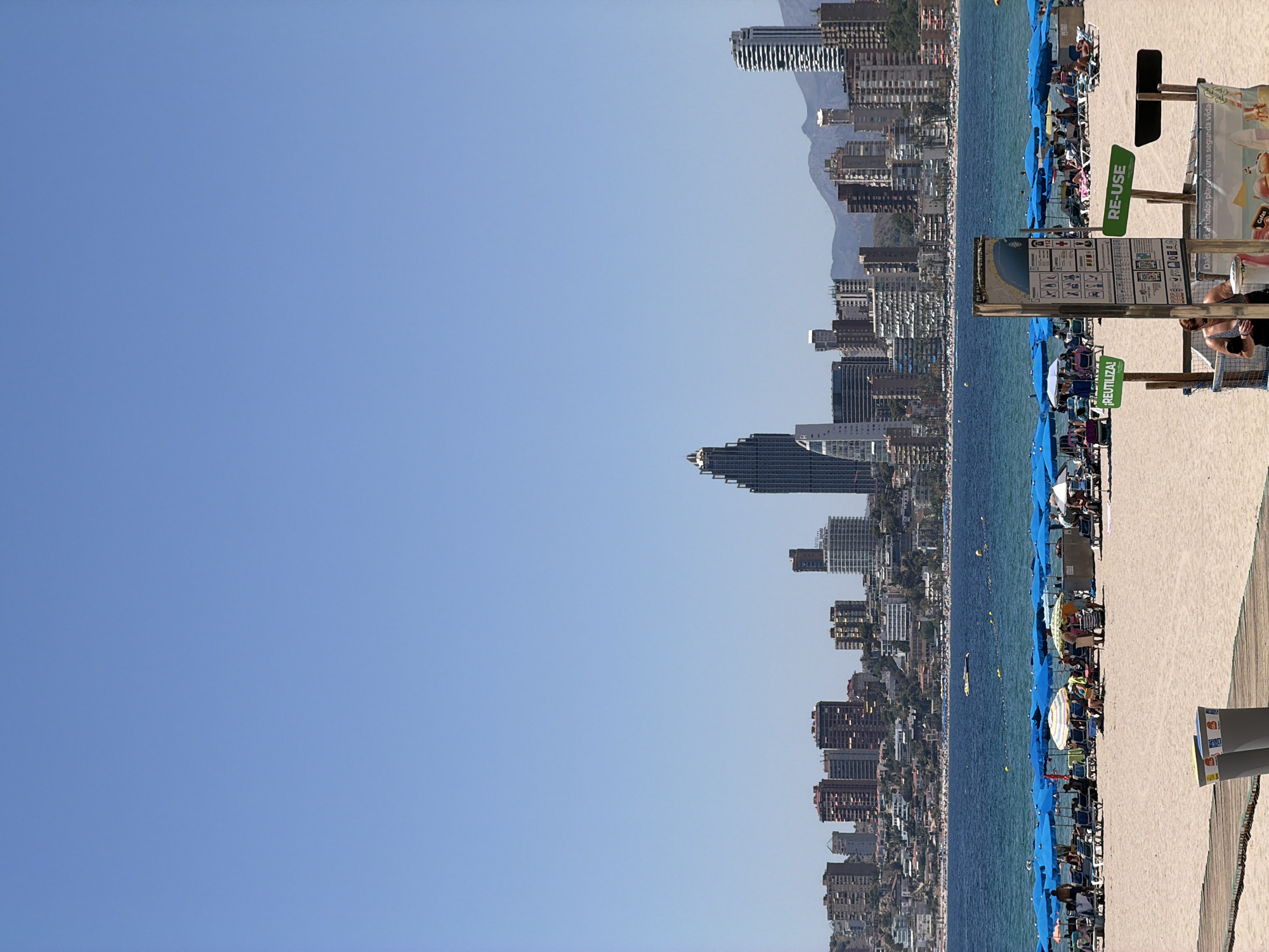 The image depicts a sunny day at a crowded beach in Benidorm, Spain, with the city's iconic skyscrapers forming a backdrop. In the foreground, a wooden boardwalk leads towards the sand, where two grey trash receptacles are visible. Beyond the boardwalk, beachgoers are scattered across the sand, many relaxing under a multitude of blue umbrellas. Some individuals are seated on lounge chairs, while others are standing and conversing. A series of beach umbrellas are lined up, creating a vibrant, colorful display.
Further back, the turquoise waters of the Mediterranean Sea stretch towards the horizon, dotted with a few small boats and buoys. The city of Benidorm itself rises from the coastline, a dense collection of modern high-rise buildings and apartment complexes. Notably, one particularly tall, dark skyscraper with a distinctive architectural design stands out amongst the others. In the distance, the rolling hills of the surrounding landscape are visible, partially obscured by the haze of the warm weather. A sign with instructions for "RE-USE" and recycling is positioned on the beach, suggesting an effort towards environmental awareness. The overall scene is one of a bustling tourist destination, characterized by its beach, sea, and urban architecture.