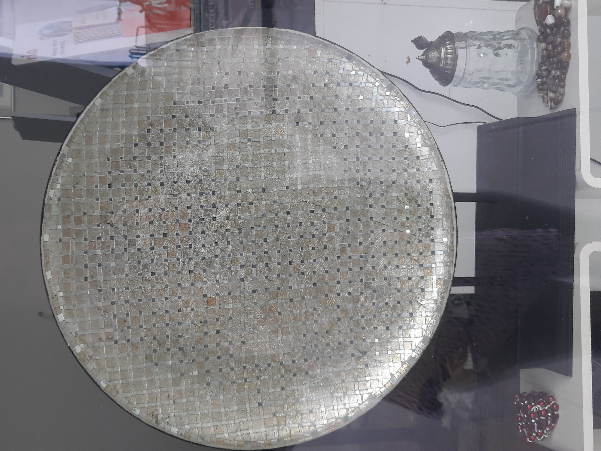 The image displays a large, round mosaic table surface with a pattern of small square tiles in alternating light and dark shades, arranged in a grid. A portion of the table is visible, suggesting it is a centerpiece in a display. To the right of the table, a clear glass jar with a silver lid is present, accompanied by a strand of beads. To the left of the table, another strand of beads, this time dark red and silver, is visible. The background appears to be a shop window, with reflections of the interior and possibly some signage visible on the right. The overall impression is a display of decorative items, likely within a retail environment. Without further information, the specific location in Rosenheim, Germany, cannot be determined from the visual content alone.