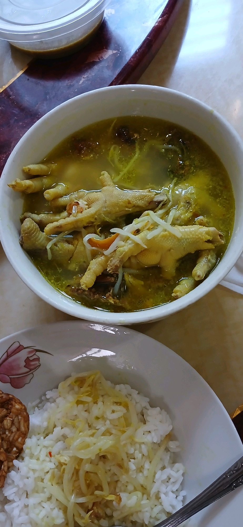 In Tanjung Pinang, Indonesia, a close-up, overhead shot shows a white bowl filled with a savory broth containing chicken feet, bean sprouts, and what appears to be small pieces of dried shrimp or spices. Adjacent to the bowl of soup, a white plate holds a serving of white rice topped with more bean sprouts and a piece of fried tempeh on the left side. The food is arranged on a table with a marble-like pattern and a dark brown trim. A clear plastic container with a lid, holding a yellowish liquid, is visible in the upper left corner, suggesting it might contain a dipping sauce or another beverage. The lighting suggests an indoor setting, possibly a home or a local eatery. The composition is focused on the food, highlighting the textures and colors of the dishes.