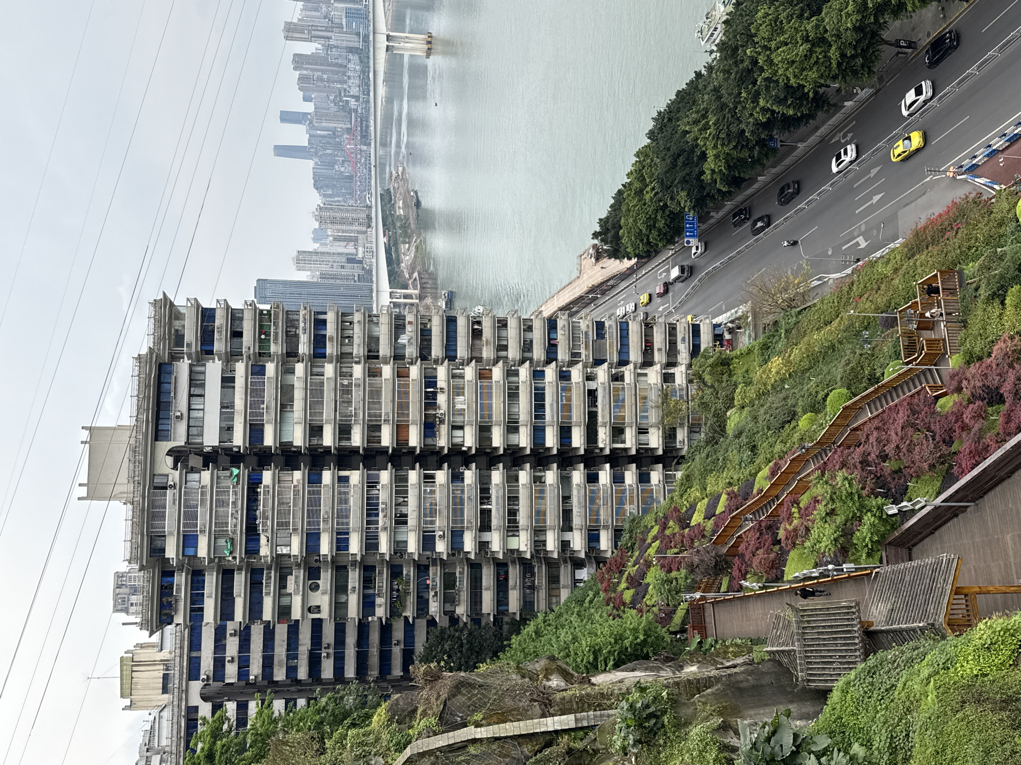 The image captures a view in Yuzhong District, China, presenting a juxtaposition of urban infrastructure and natural elements. Dominating the foreground is a multi-story residential building with a repetitive facade pattern of windows and balconies, indicating a high-density urban dwelling. To the right of the building is a steep, verdant hillside with terraced pathways and vegetation, contrasting with the building's rigid structure. A road runs along the hillside's edge, with moving vehicles of various colors. In the background, across a wide river, a dense cityscape is visible under an overcast sky. Overhead power lines run parallel to the building's height. The image suggests the intersection of modern urban life with elements of the natural landscape in a bustling Chinese city.