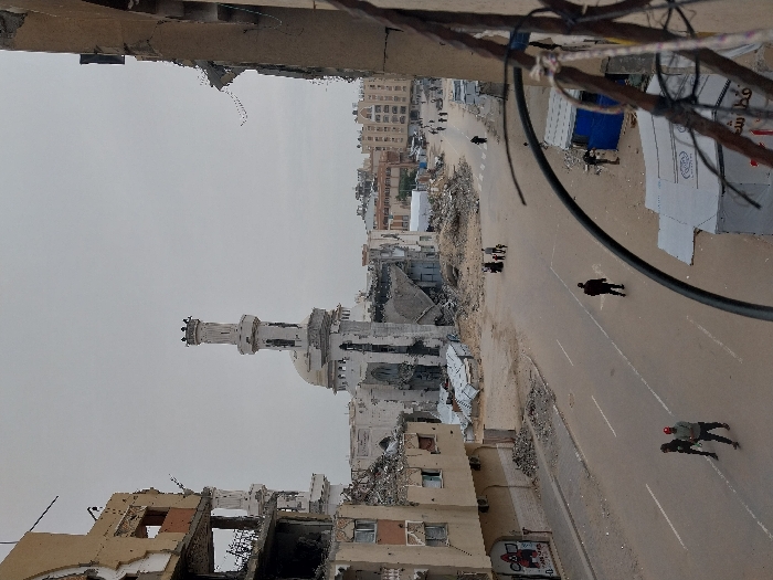 The image presents a devastated urban streetscape in Khan Yunis, Palestinian Territory, under an overcast, light grey sky. In the mid-ground, a prominent mosque with a tall minaret and a dome exhibits extensive structural damage, surrounded by significant piles of concrete rubble and debris. To the left, a multi-story building, light brown in color, is severely damaged; large sections of its external walls are missing, revealing exposed interiors and skeletal rebar. Rubble is piled at its base. The immediate right foreground shows the remains of another damaged building with a broken arched opening and exposed rebar and wiring.
An asphalt road, marked with faded white lines, traverses the scene, appearing devoid of vehicles. Numerous individuals are visible on foot: two men walk from right to left in the foreground; a lone man walks away from the camera in the center of the road; a group of approximately five individuals, including adults and children, walks on the right side of the road further in the distance; and several more individuals are scattered in the background. A small blue and white makeshift shelter stands by the roadside on the right, adjacent to a white tarpaulin covering materials on the ground, featuring visible Arabic text. The environment is characterized by widespread destruction and rubble, indicating extensive damage to infrastructure.