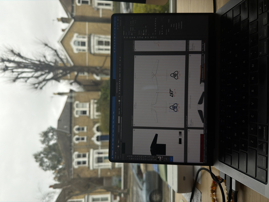 The image presents an indoor perspective looking out through a window onto a residential street in London, United Kingdom.
In the foreground, a silver-grey MacBook Pro laptop is open on a light wooden surface. The laptop screen displays design software, likely for apparel, showing multiple views of garment mock-ups. On the left side of the screen is a flat black long-sleeved top. The central area features schematic diagrams of a garment, possibly a jacket or sweatshirt, from front and back perspectives, complete with measurement lines and three logos: two stylized interlocking triple-loop designs, and a text logo "LST" in red and black. Further garment components are visible in the lower section of the screen. A black power cable with a USB-C connector is plugged into the laptop's left side, running towards a light-colored rectangular power adapter or external device situated on the wooden surface. A string of amber-colored spherical beads rests near the cable.
The background, visible through the window, depicts an overcast sky above a street lined with traditional British houses. These buildings are constructed of light brown/yellow brick, featuring white-framed sash windows and prominent bay windows on multiple floors. Dark roofs and visible chimneys are present. The central house directly opposite has a dark blue front door. Several deciduous trees, bare of leaves, are scattered along the street, with a particularly tall, pruned tree prominent on the right side. A light-colored car, possibly silver or light blue, is parked on the street to the left. No individuals are visible in the scene. The overall environment suggests a typical residential area within Greater London.
