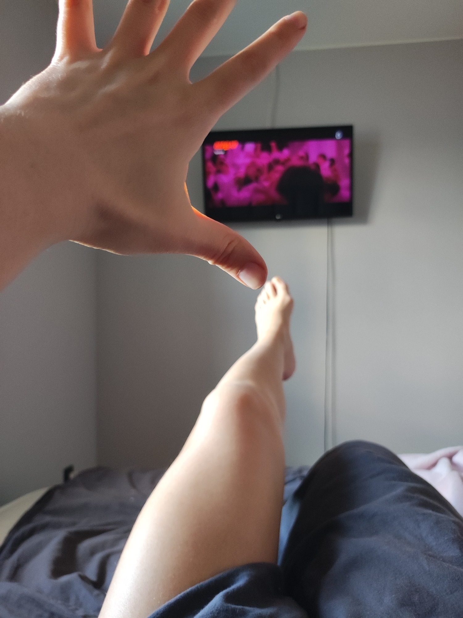 This media file captures an indoor scene, likely a bedroom or living room, with a focus on a person relaxing. The setting features light gray walls.
The primary subjects are parts of a person's body: a hand, a leg, and a foot. In the foreground, a left hand is held up towards the camera, fingers spread, with light illuminating the back of the hand. The light appears to be coming from the left, possibly a window. Further back, a bare leg is extended, with the foot slightly raised. A portion of another leg or torso, covered by dark fabric, is visible in the bottom right corner, suggesting the person is lying down on a bed or sofa. Dark gray bedding is also visible in the lower-left foreground.
The main activity depicted is one of relaxation, with the person seemingly watching television. A small, black flat-screen television is mounted on the wall opposite the viewer, towards which the person's leg is extended. The TV screen displays vibrant pink and purple hues with dark, blurred silhouettes, suggesting a performance, concert, or party scene is being watched. A single white cable runs vertically down the wall from the TV. The hand gesture in the foreground, with fingers spread, might be a playful use of forced perspective.
Notable details include the bright lighting, suggesting it is daytime. The scene appears casual and comfortable. There is no discernible text visible on the television screen or elsewhere in the image. This indoor scene is situated in Votkinsk, Russia.
