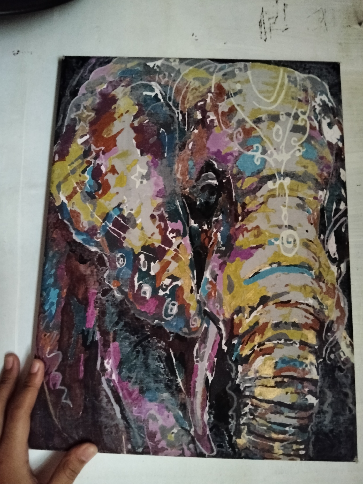 The image displays a close-up, slightly angled view of a vibrant, abstract painting of an elephant. The artwork is rendered in a bold, expressive style with a rich palette of colors including yellows, blues, pinks, purples, browns, and grays, applied with visible brushstrokes. Decorative white patterns, resembling swirls and geometric shapes, are superimposed on the elephant's trunk and forehead. The background is dark and indistinct, focusing all attention on the textured, colorful subject. A person's hand is visible in the lower left corner, appearing to hold or steady the painting, with fingers gently touching its surface. The painting is presented against a plain, off-white background, possibly a wall or a flat surface, with some faint, scribbled markings visible in the upper background area. There is no identifiable information within the image to suggest a location in Davao City, Philippines.