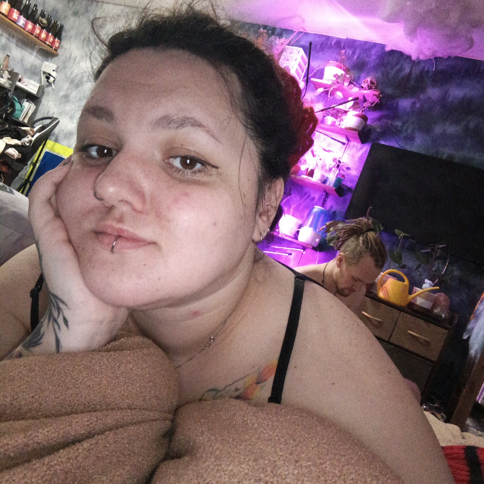 A woman with brown hair pulled back in a bun and a lip piercing rests her head on her hand, looking directly at the camera. She wears a black-strapped garment and a necklace. Tattoos are visible on her hand and chest. Behind her, a man with dreadlocks tied up is bent over a dresser on which sits a yellow watering can, plants, and other miscellaneous items. Shelves with plants, illuminated by a pink light, are mounted on the wall above the dresser. A dark flat-screen TV is also visible on the wall. A shelf with bottles is partially visible on the left. The location could not be determined based solely on the image.