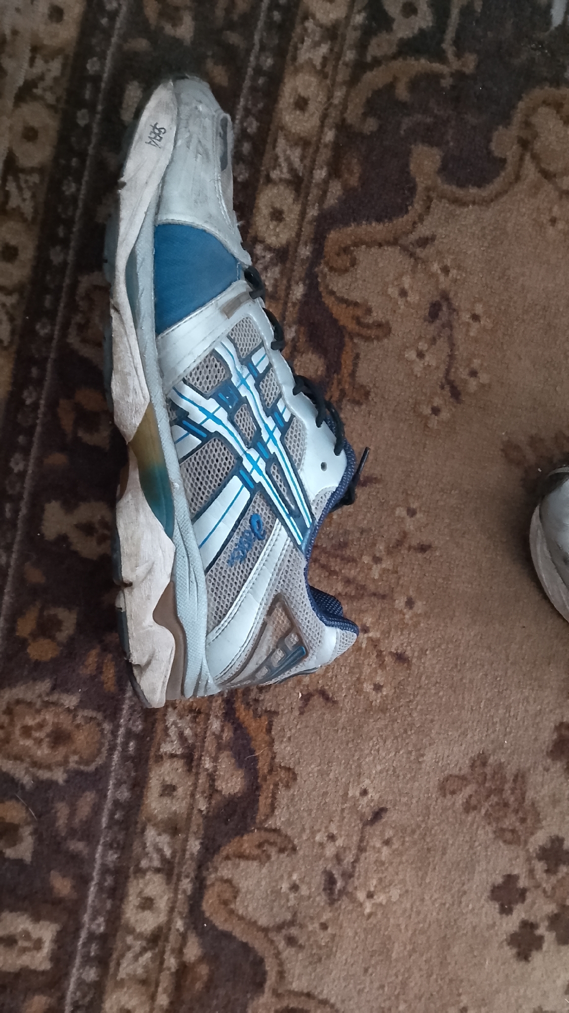 The image shows a single ASICS running shoe, primarily white and blue, lying on its side on a brown carpet with a floral pattern. The shoe appears worn and dirty, with some scuff marks on the sole and upper. The ASICS logo is visible on the side of the shoe. The carpet is textured and appears to be in a room. There is another white shoe visible in the upper right corner of the image, also on the carpet. The image context places this scene in the city of Ustyvytsia, Ukraine.