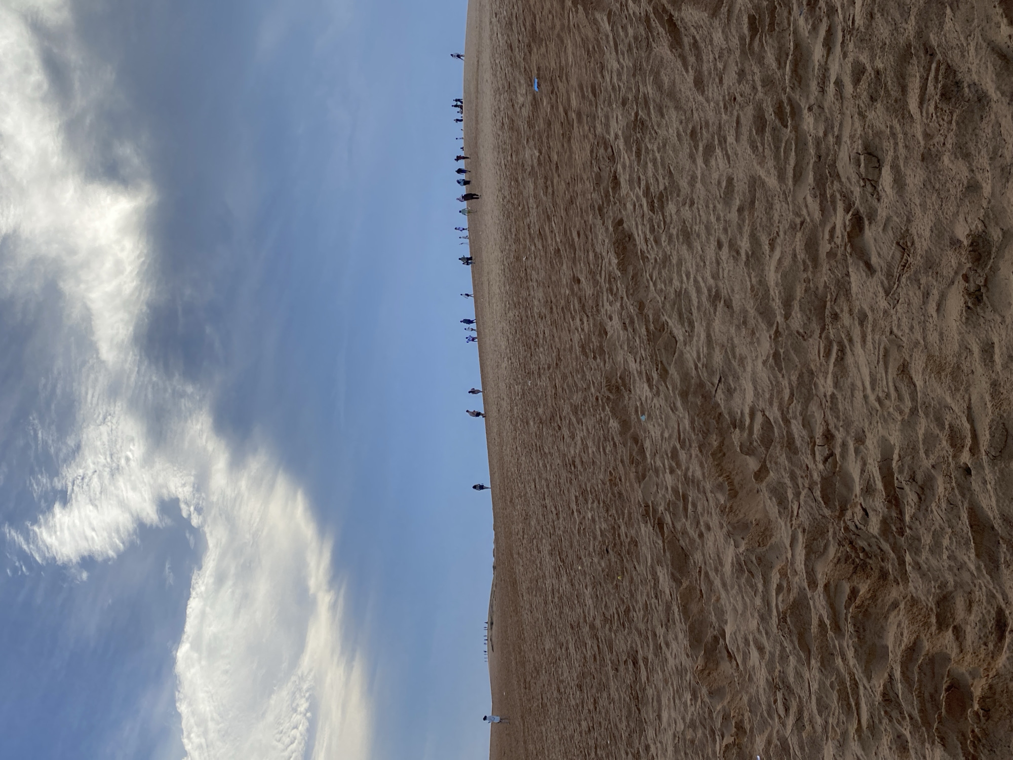 The image captures a vast landscape featuring a large sand dune under an expansive sky. The perspective suggests a view looking along the side of a dune, with the sandy expanse dominating the lower right portion of the frame. A distinct horizon line separates the earth from the sky.
A long line of small figures, identified as people, are seen walking along the crest of the sand dune, appearing as silhouettes against the brighter sky. They are spaced out, forming a trail that stretches into the distance. The sand dune itself exhibits detailed texture, with ripples and undulations formed by wind, and visible tracks from previous activity.
The upper left portion of the image is filled with a bright blue sky, adorned with scattered white and light grey clouds. Some clouds are wispy and elongated, while others are denser and more patchy. The illumination suggests either early morning or late afternoon light, casting a soft glow across the scene without harsh shadows.
The activity taking place is people trekking or walking along the ridge of a sand dune, likely for sightseeing or recreation. The scene conveys a sense of scale and the natural beauty of a desert or coastal dune environment. This setting aligns with the provided location context of Mũi Né, Vietnam, known for its significant sand dunes. No discernible text is visible within the image.