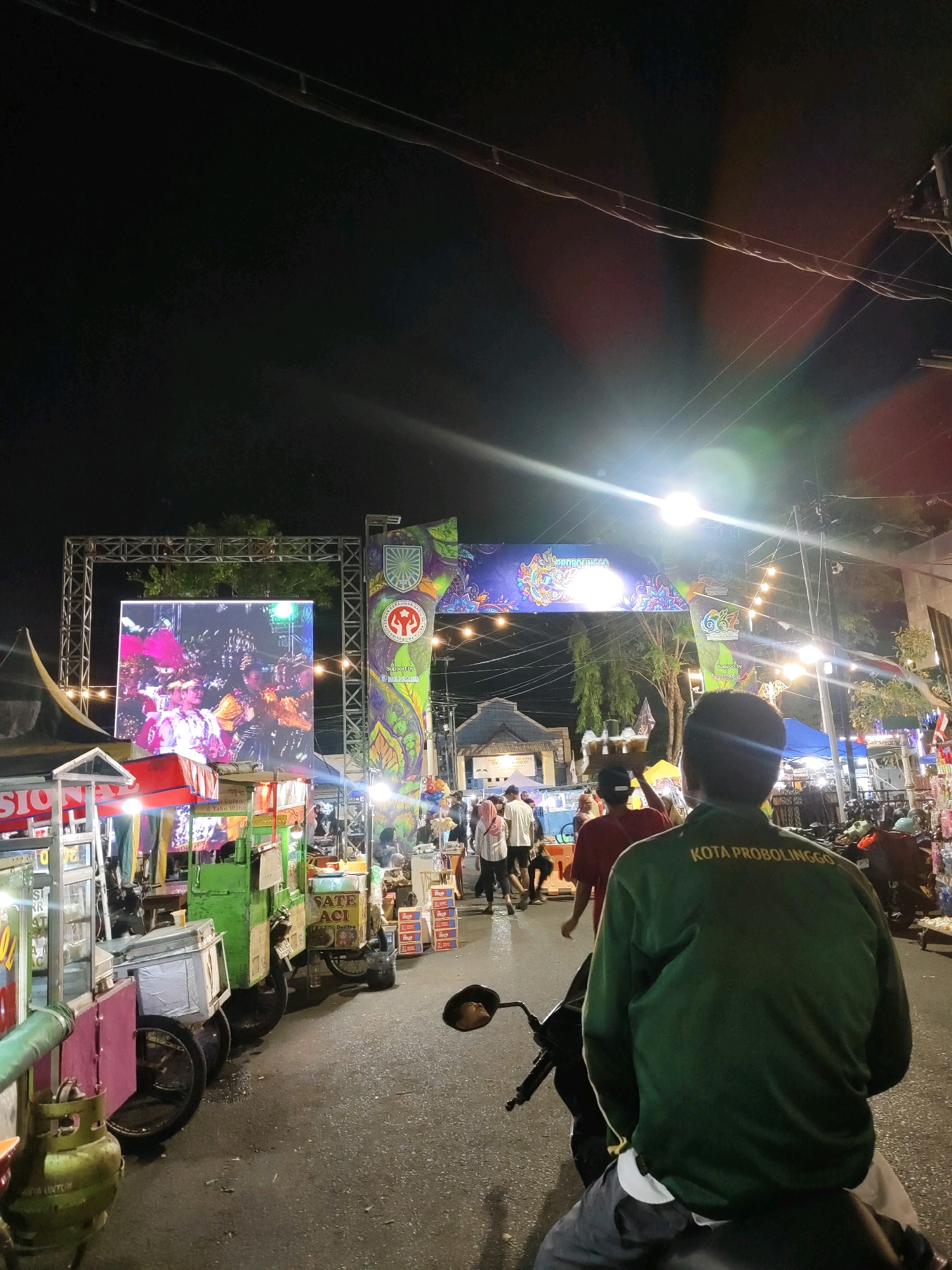 The image depicts an outdoor night scene in the city of Probolinggo, Indonesia, likely at a festival or market. In the foreground, a person is seated on a motorcycle, facing away from the camera. They are wearing a dark green jacket with yellow text on the back that reads "KOTA PROBOLINGGO".
To the left of the motorcycle, a row of food stalls or vendors are set up. These stalls are brightly lit and feature various goods, including what appear to be food items in glass cases and canisters. One stall has a prominent display of gas cylinders.
Further into the scene, an arched structure spans across the street, adorned with colorful banners and illuminated by string lights. Behind this arch, a large LED screen displays an image of people in traditional costumes, suggesting a cultural performance or display. Other stalls and a crowd of people are visible in the background, indicating a lively public gathering. The overall environment is a street scene at night, illuminated by artificial lights from the stalls, banners, and the screen.