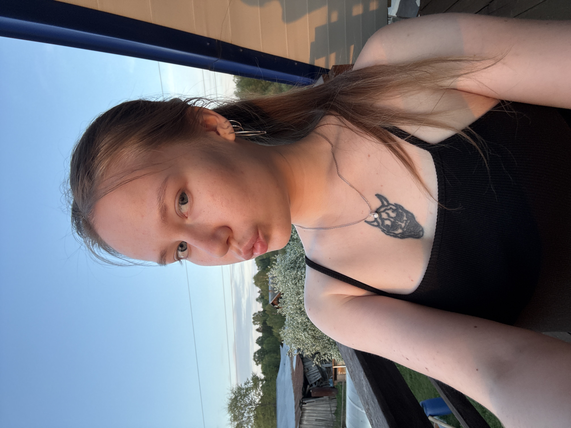 A selfie of a light-skinned young woman, facing left with her head tilted, is set against an outdoor background, likely in a rural area. She has brown hair and is wearing a black tank top and silver necklace with a wolf-like pendant. Silver earrings are visible in her left ear. The background features trees, a few rural-looking buildings, and a sky transitioning from blue to a lighter shade near the horizon, suggesting it may be evening. The photo is taken from an elevated position, possibly a porch, with a railing visible in the lower right corner. Location: Unknown, Russia.