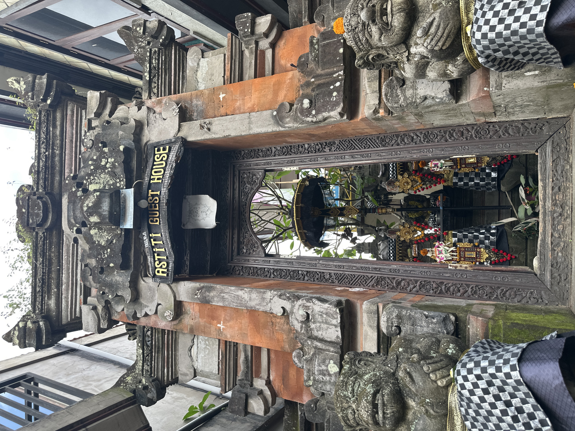 A stone entrance gate with intricate carvings and statues of mythical guardians stands in front of the Astiti Guest House in Ubud, Indonesia. The gate is adorned with traditional Balinese architectural elements, including ornate stone carvings and terracotta bricks. The statues, positioned on either side of the entrance, appear to be weathered and covered in moss, suggesting they are ancient. They are dressed in checkered cloths. The entrance leads into a courtyard with lush greenery and a decorated altar. The overall scene conveys a sense of cultural richness and tranquility, characteristic of the Ubud district.